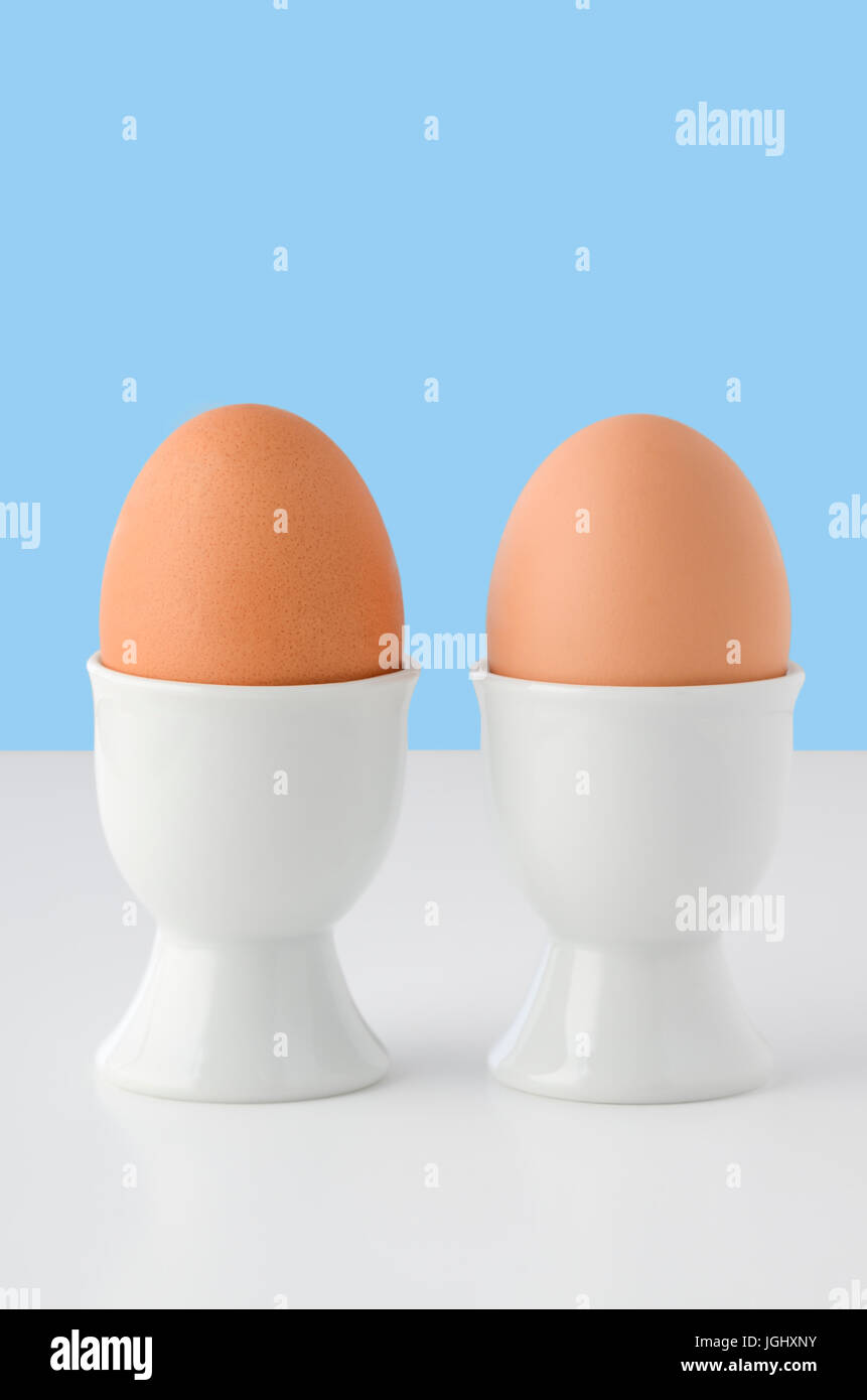 Deux oeufs brun en plain white porcelain egg cups sur tableau blanc avec la lumière, fond bleu lumineux. Espace copie ci-dessus. Banque D'Images