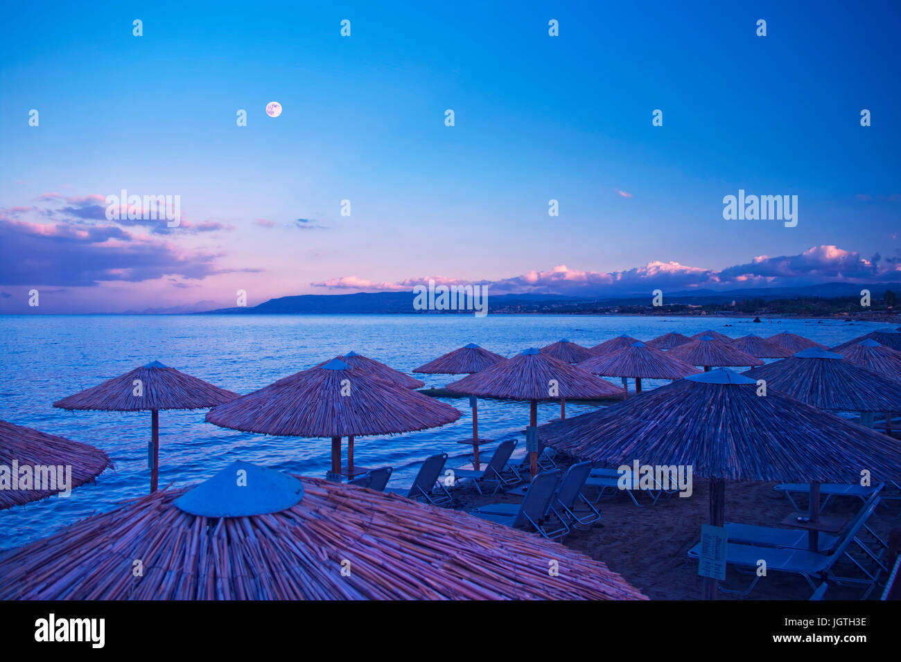 Des parasols et transats sur la plage la nuit avec les montagnes et la pleine lune en arrière-plan à Georgioupolis, Crète, Grèce Banque D'Images