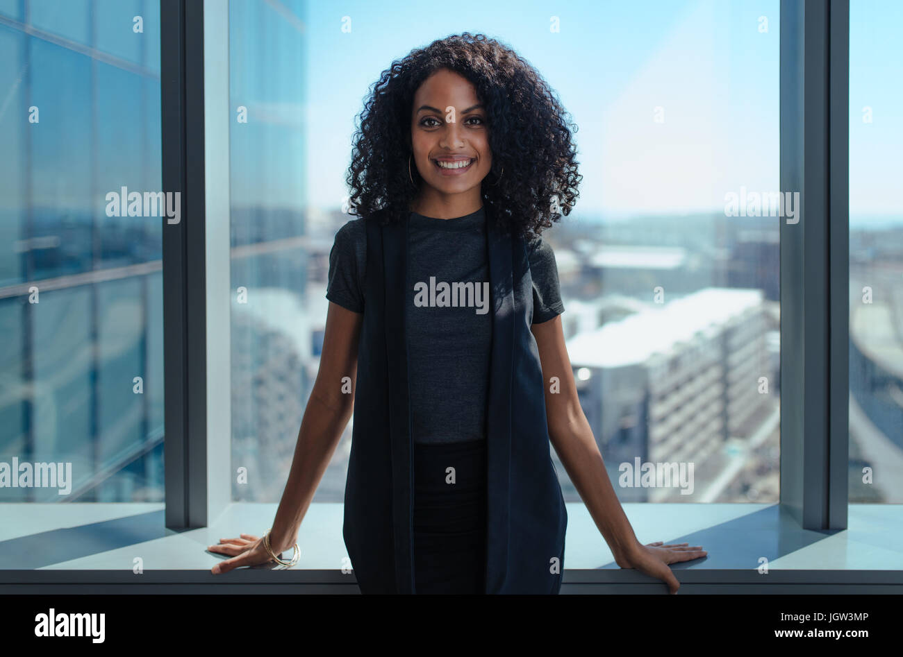 Smiling businesswoman standing dans son bureau dans un immeuble donnant sur le paysage urbain. Femme aux cheveux bouclés, debout près de fenêtre dans Office. Banque D'Images