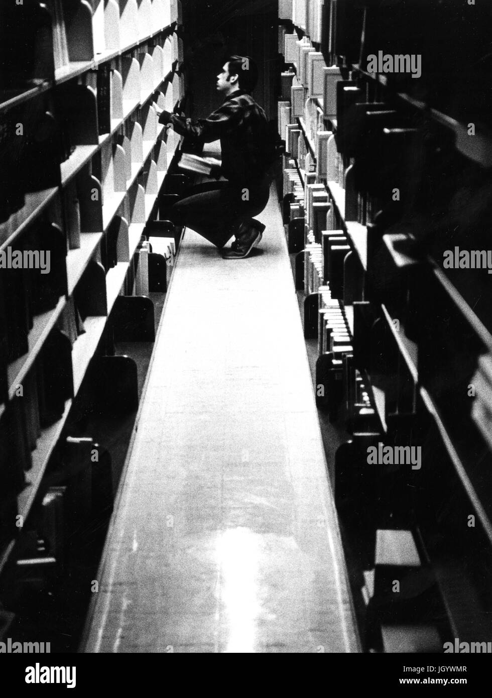 Candid portrait d'un étudiant de l'Université Johns Hopkins crouching dans les rayons à la recherche de livres à l'Université bibliothèque Milton S. Eisenhower, à Baltimore, Maryland, 1965. Banque D'Images