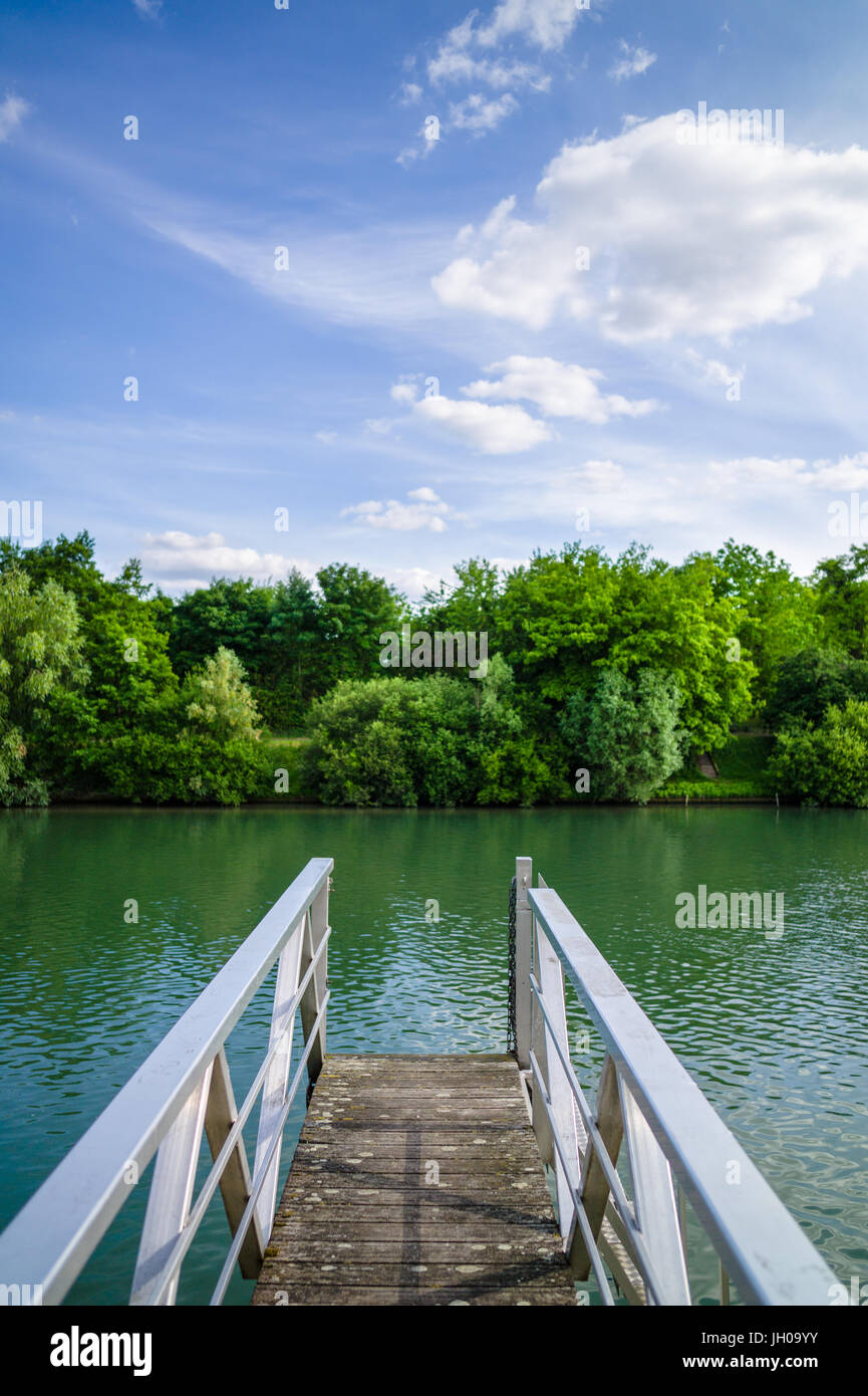 Un ponton en bois avec rampe métallique sur une rivière avec des arbres et de la végétation sous un ciel bleu avec des nuages blancs. Banque D'Images
