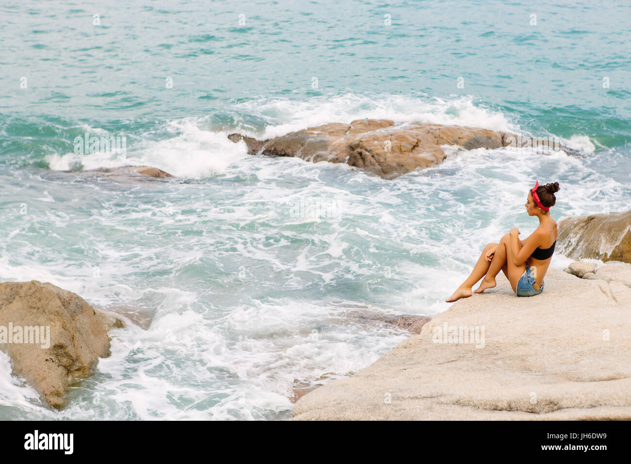 Belle fille de short court en denim rouge, bandeau et soutien-gorge noir assis sur grosse pierre sur la plage en mer océan tempête. Elle toucher son long monter les jambes. Co Banque D'Images