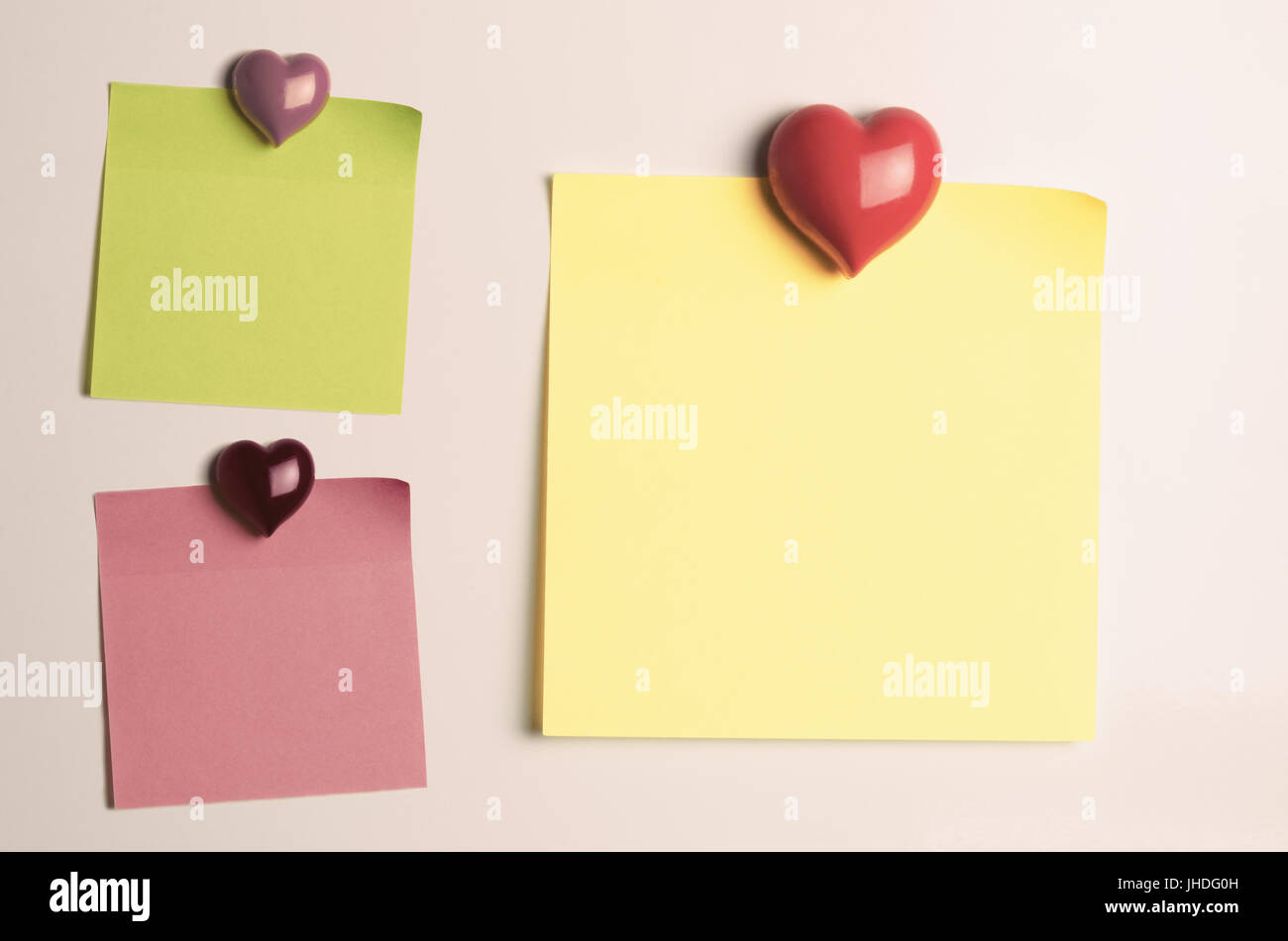 Sticky note trois carrés dans du papier vert, rose et jaune, coincé à porte du frigo avec des aimants en forme de coeur. Laissée vierge pour fournir une copie de l'espace. Banque D'Images