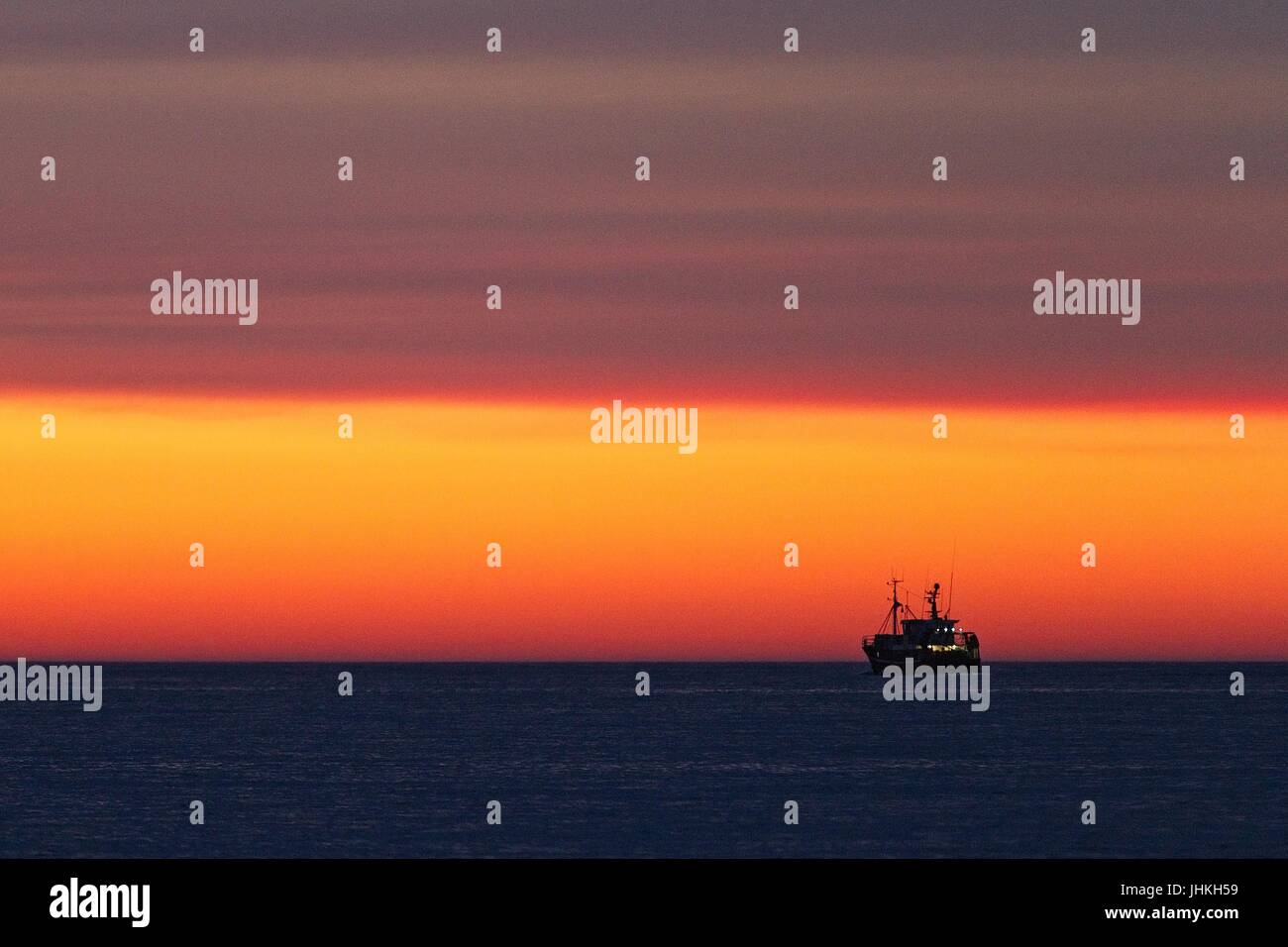 Silhouette d'un bateau de pêche sur la mer juste après le coucher du soleil. Couleur rouge jaune doré. Ciel avec nuages. Quelques lumières sur le bateau de pêche. Banque D'Images