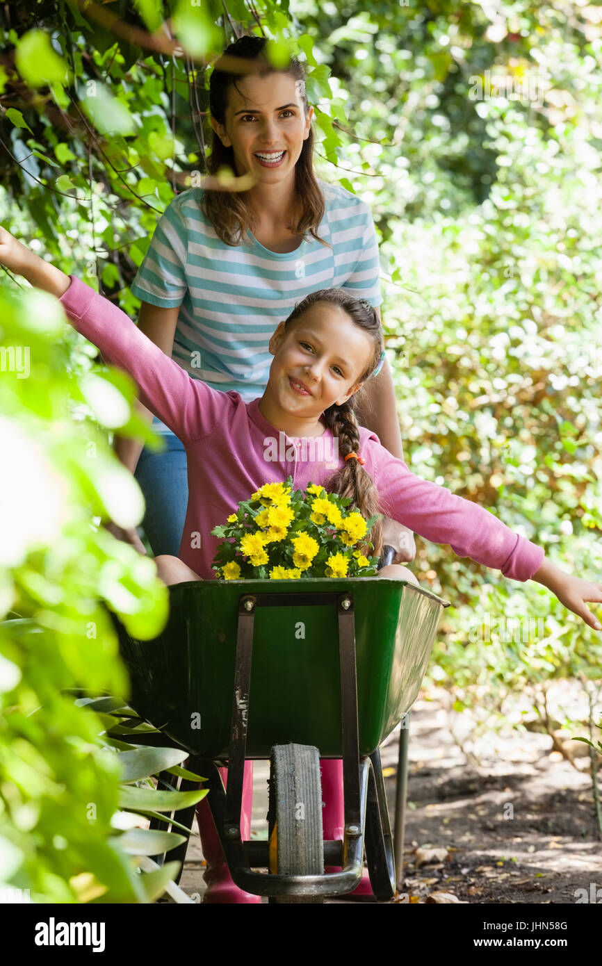 Girl enjoying with arms outstretched while smiling mother pushing wheelbarrow sur sentier par les plantes Banque D'Images