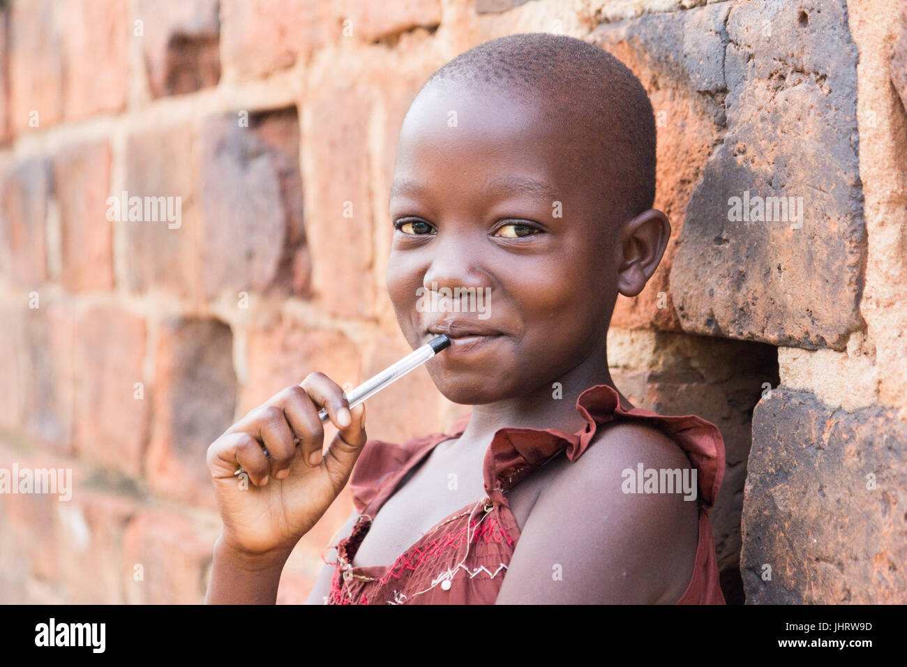Un noir de 11 ans jeune fille ougandaise en souriant et tenant un stylo contre sa bouche et appuyé contre un mur de briques à l'appareil photo Banque D'Images