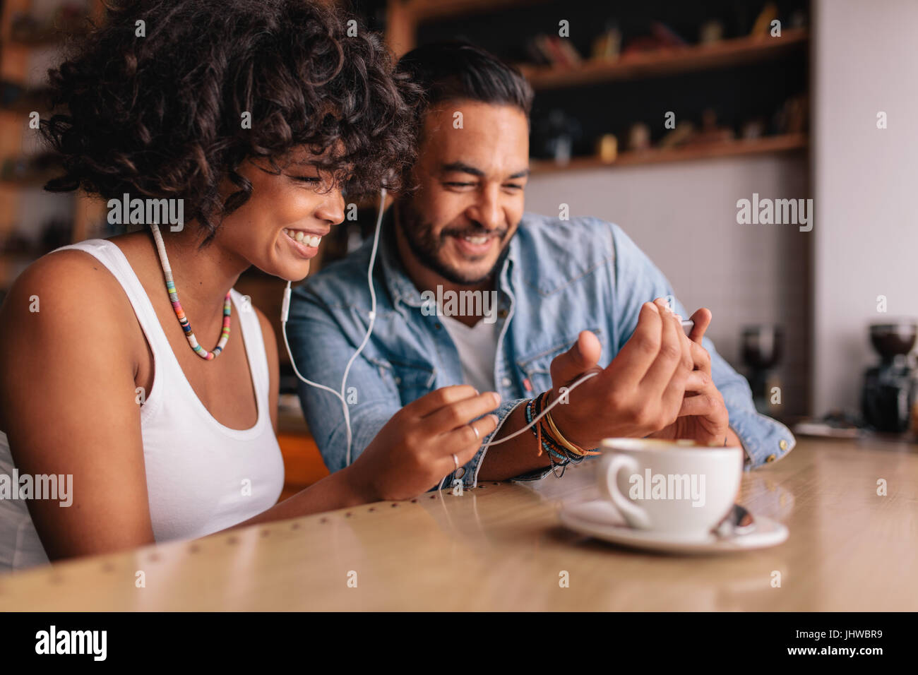 Happy young couple sitting at coffee shop chat vidéo sur téléphone mobile. Jeune homme et femme avec des écouteurs au café à l'aide de smart phone. Banque D'Images