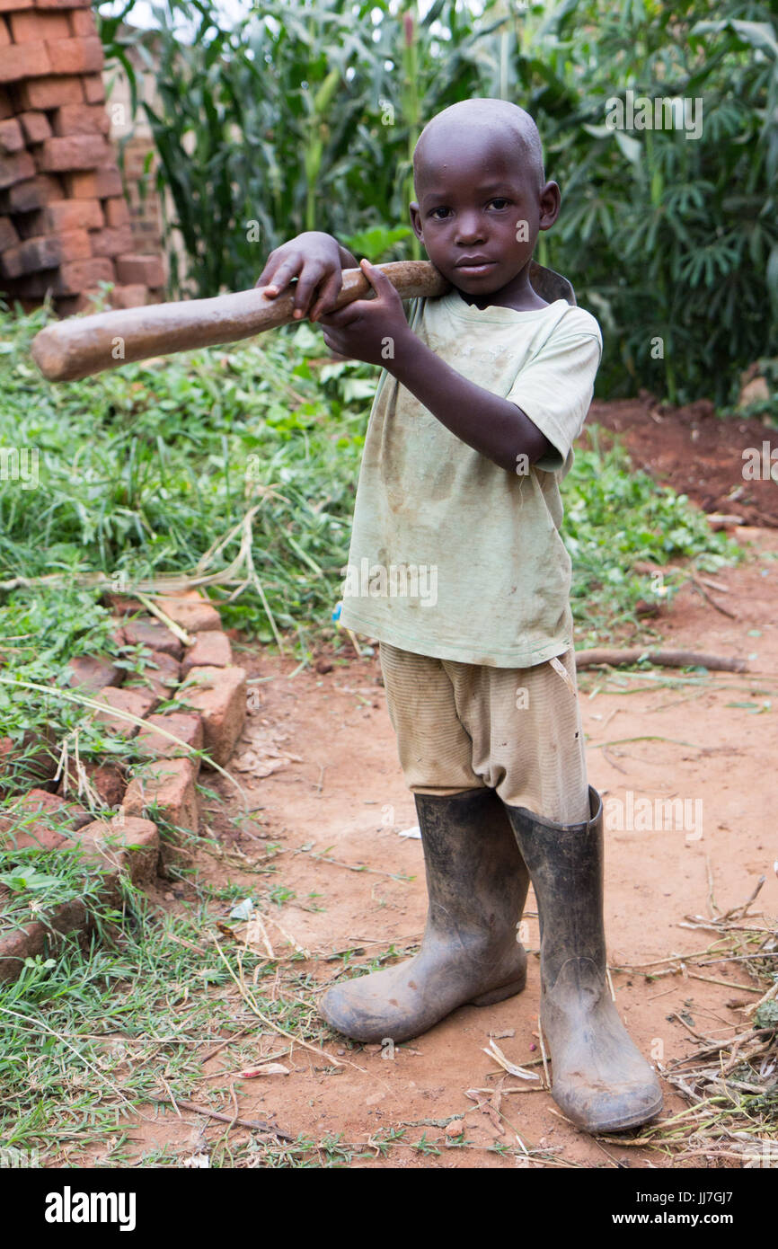 Un enfant ougandais africaine noire garçon tenant une pioche sur son épaule. Le garçon est en mauvais rag vêtements et bottes de caoutchouc Banque D'Images