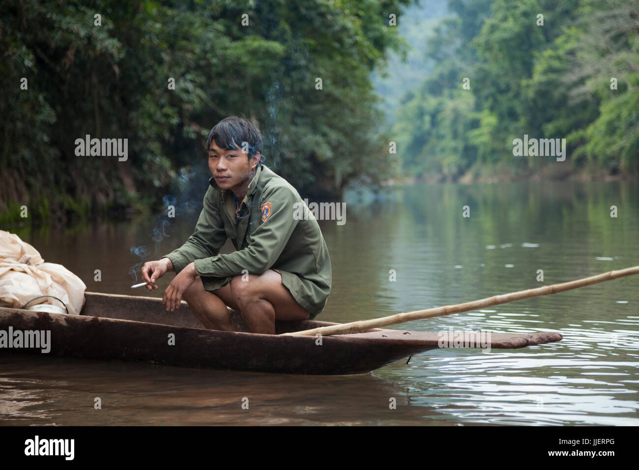 Un jeune homme sur une expédition de chasse et de pêche fume une cigarette dans son étang bateau sur la rivière Nam Ou, au Laos. Banque D'Images