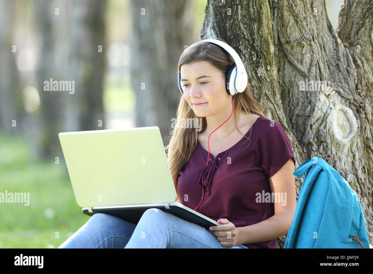 Seule fille de l'adolescence l'étude regardant la vidéo tutoriaux en ligne dans un ordinateur portable assis sur l'herbe s'appuyant sur un arbre dans un parc Banque D'Images