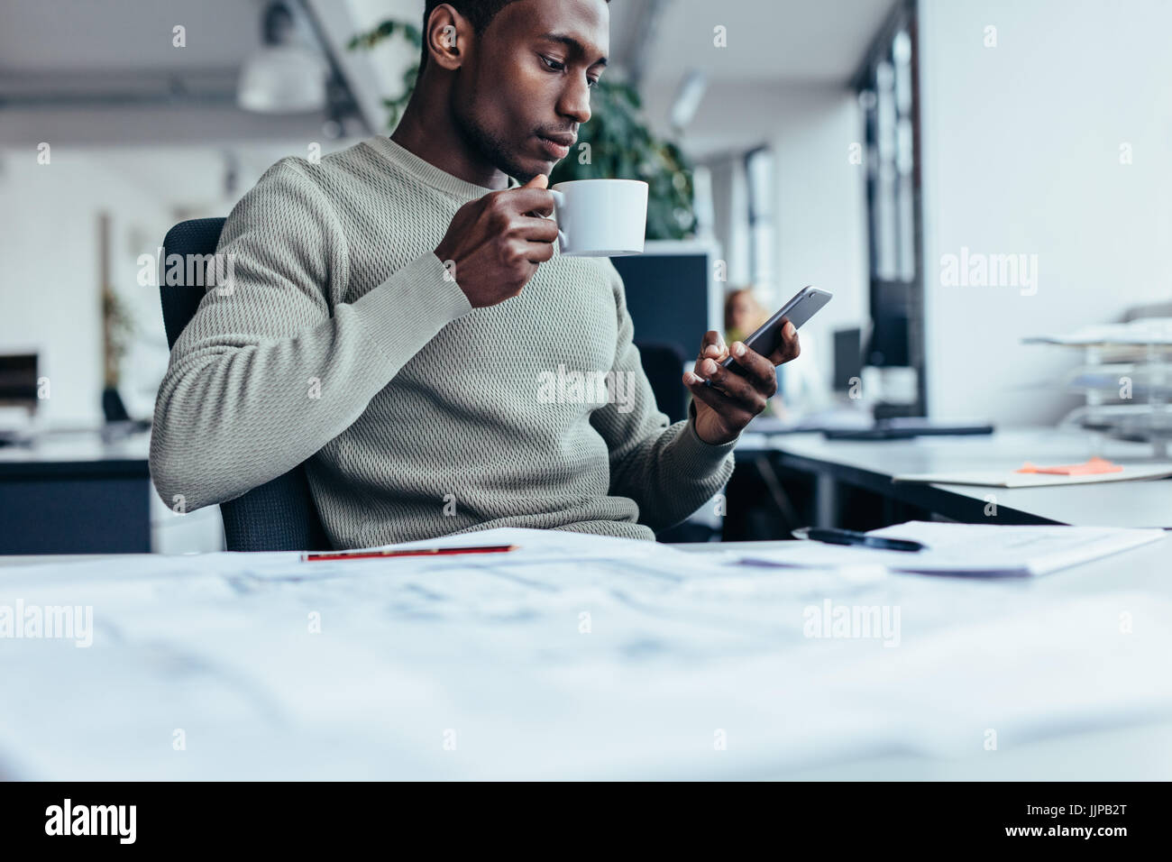 Businessman sitting at office desk en buvant un café. Jeune homme africain avec tasse de café et téléphone intelligent. Banque D'Images