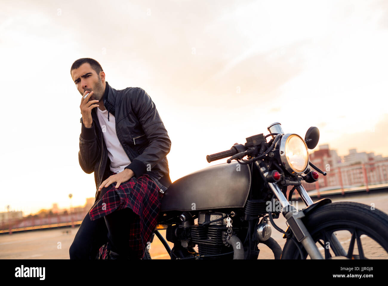 Beau rider homme avec barbe et moustache en noir veste biker fumeurs tandis que s'asseoir sur un style classique du cafe racer moto au coucher du soleil. J'ai fait sur commande de vélo Banque D'Images