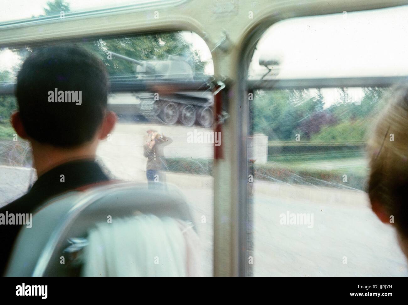 Vue de derrière d'un tourisme de l'Ouest sur un bus, regardant par la fenêtre vers la frontière entre Berlin-Est et Berlin-Ouest durant la guerre froide, avec une statue d'un tank militaire visible sur le côté est de Berlin et à l'est un soldat allemand au garde, soulevant une objection à son visage, l'Allemagne, 1970. Banque D'Images