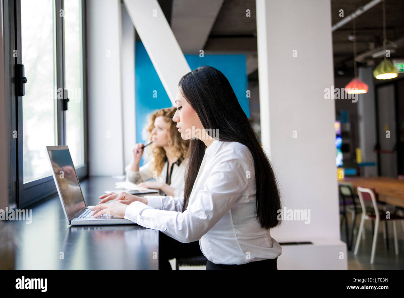 Une femme asiatique travaillant avec un ordinateur portable dans un espace de coworking. Hors foyer une femme aux cheveux blonds de Bach à regarder en dehors de la fenêtre Banque D'Images