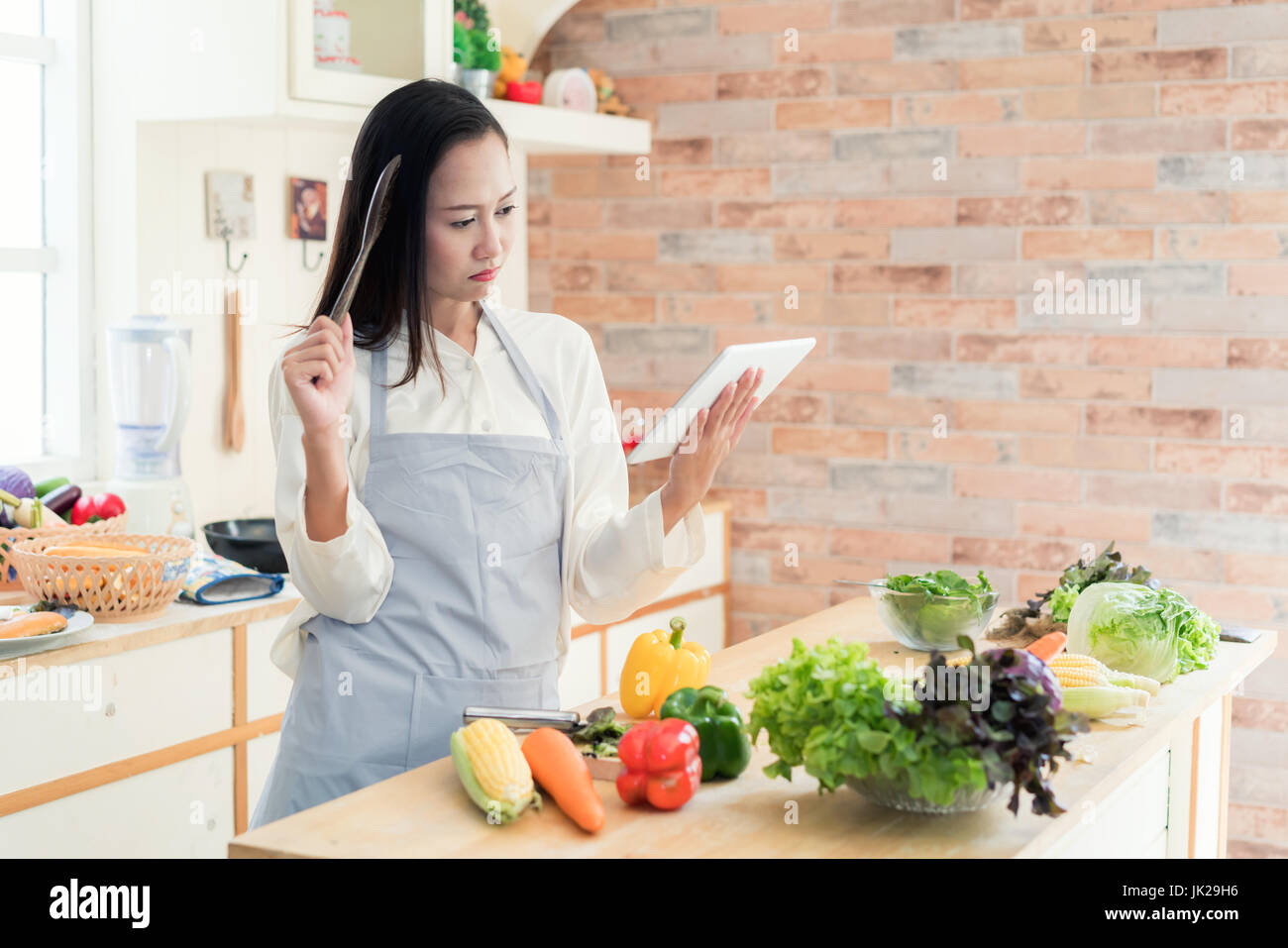 Jeune femme asiatique gaie est la cuisson dans la cuisine avec joie. Elle est debout et holding digital tablet de recette. Femme asiatique est de toucher un bois Banque D'Images