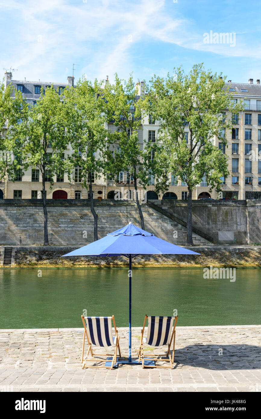 Rayé bleu et blanc deux chaises longues sous un parasol bleu sur la rive de la Seine avec arbres et immeubles parisiens typiques dans le bac Banque D'Images