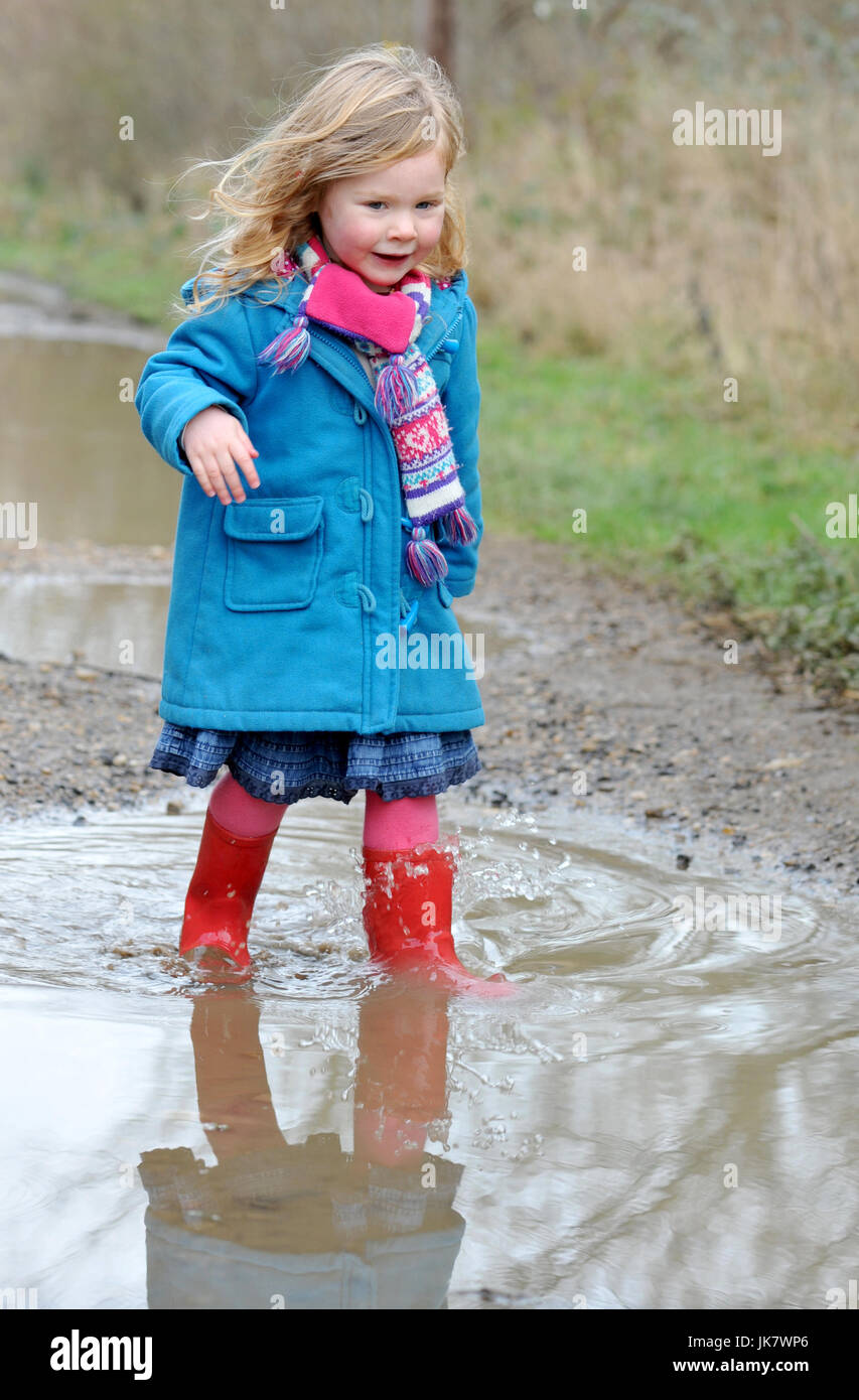 Trois ans, fille en rouge Wellington et manteau bleu patauge dans une flaque d'eau Banque D'Images