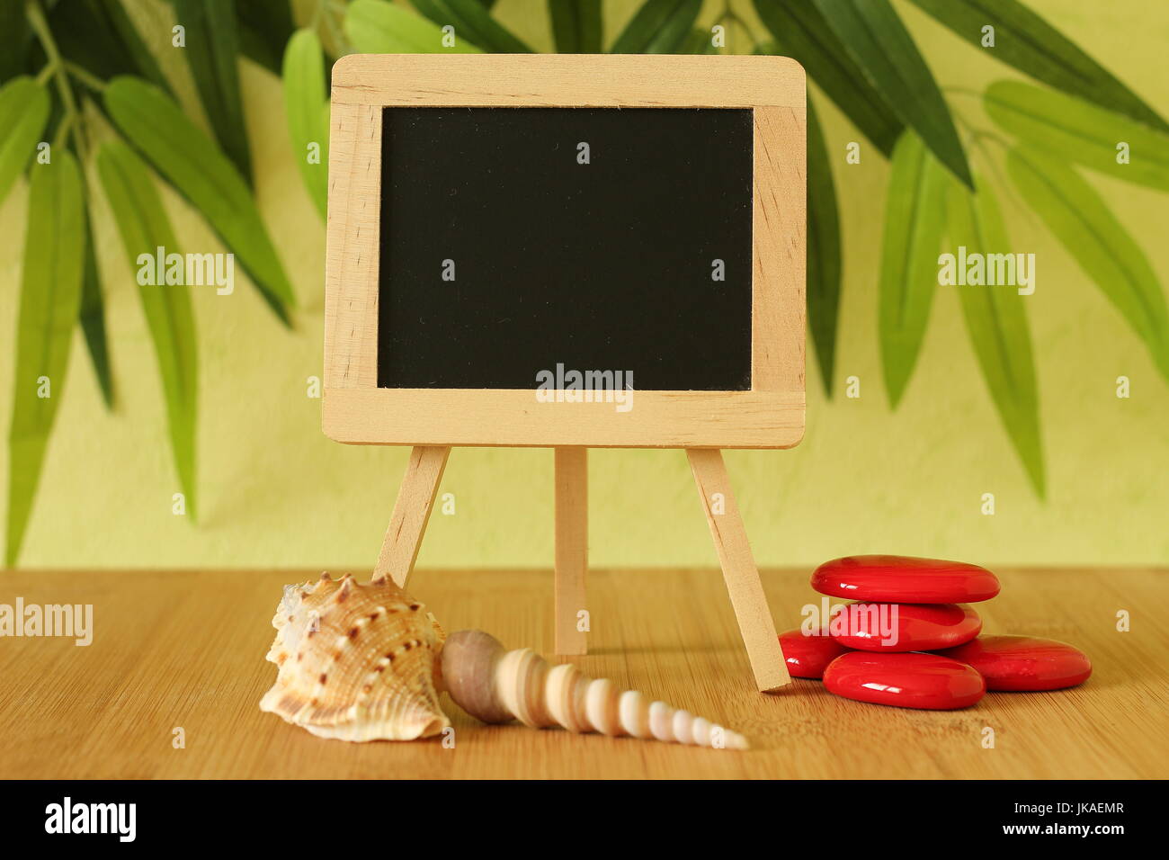 Vide petit tableau noir à écrire un message posé sur un chevalet sur un sol en bois avec plage de galets et de rouge sur un fond de feuillage vert Banque D'Images
