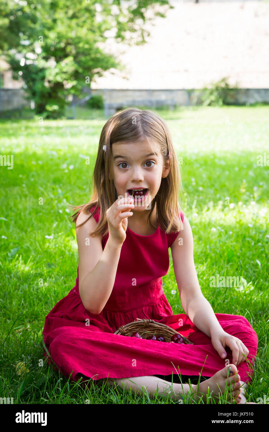 Portrait de petite fille porter du rouge robe d'assise sur un pré de manger des cerises Banque D'Images