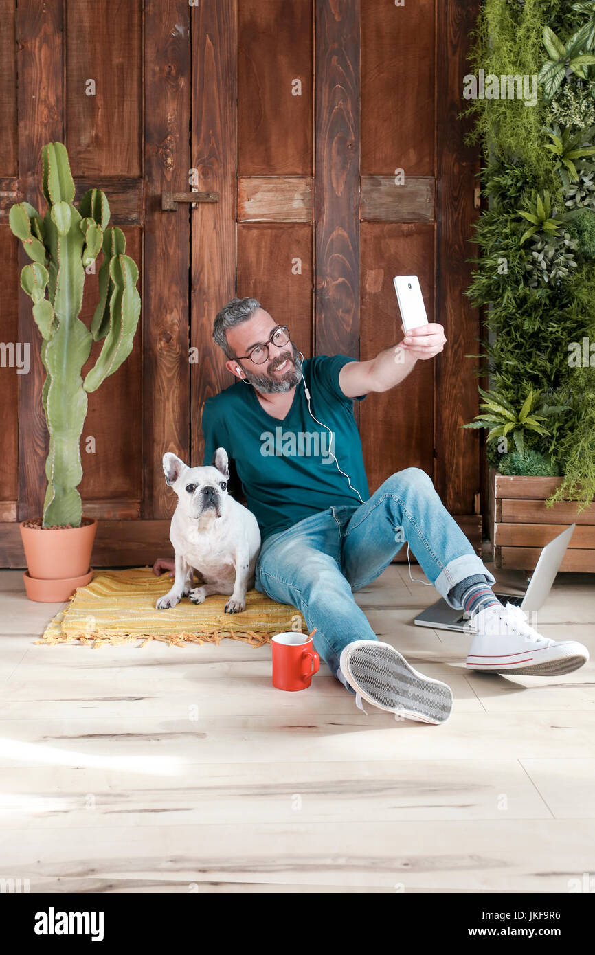 Homme barbu assis avec son chien sur le plancher à la maison à smartphone avec selfies Banque D'Images