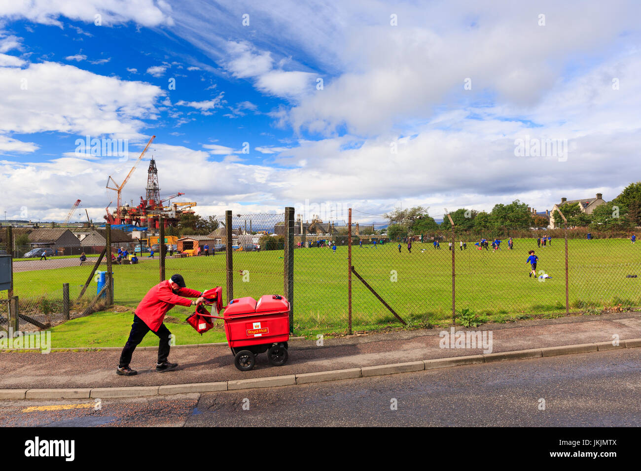 Invergordon, Écosse : facteur / factrice passant de l'école clôture aire de jeux. La plate-forme de forage en arrière-plan. Banque D'Images