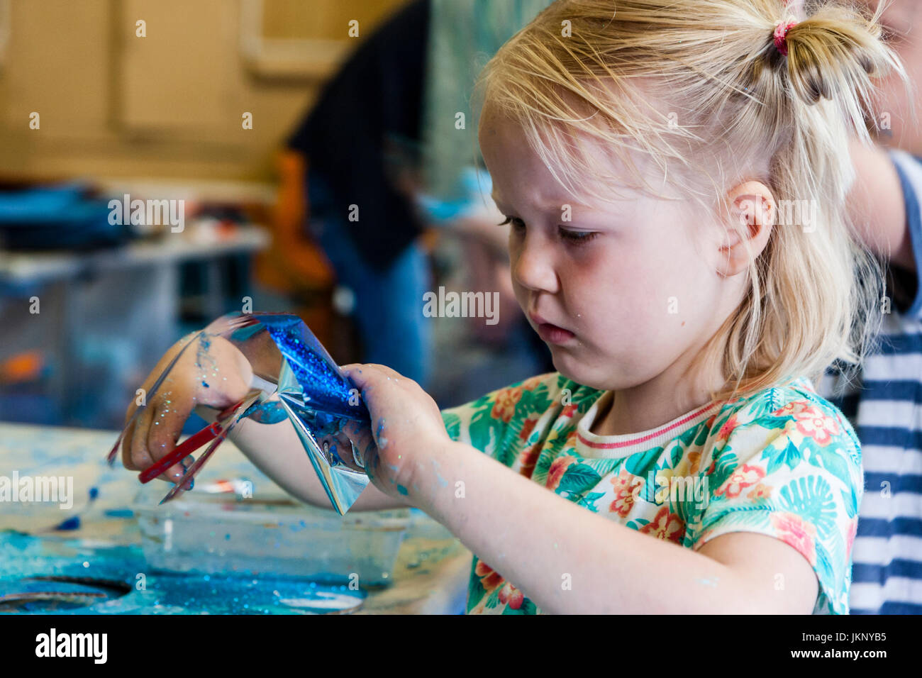 Portrait enfant blonde, fille, 5-6 ans, se concentrant très fort qu'elle tente d'utiliser des ciseaux pour couper un morceau de papier d'aluminium. Vue en gros plan, le bras et le visage. Participation à l'atelier d'art et d'artisanat. Banque D'Images