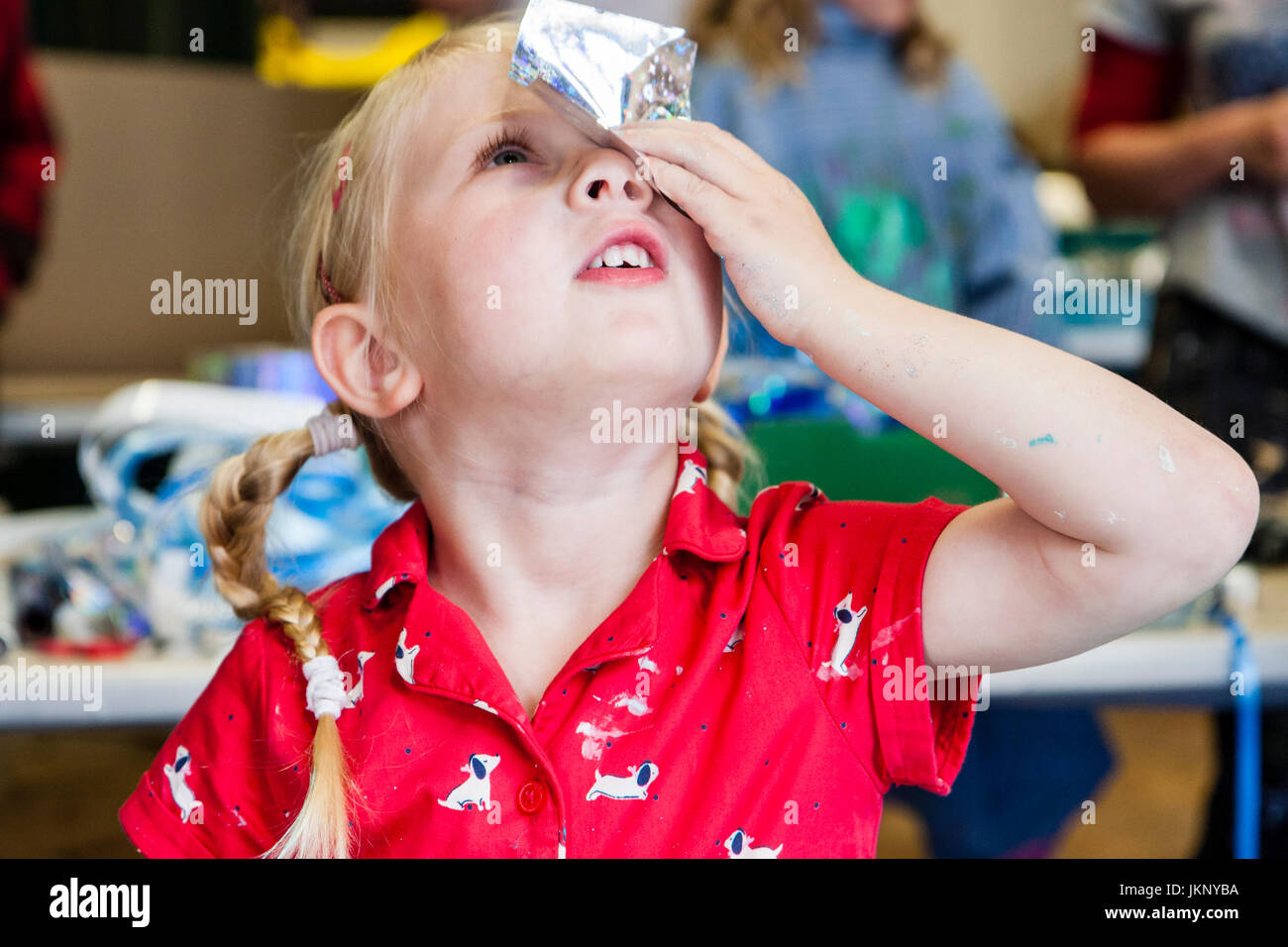 Portrait enfant blonde, fille, 5-6 ans, tenant un morceau de papier d'aluminium peint sur ses yeux pour voir si elle peut voir à travers. Vue en gros plan, le bras et le visage. Participation à l'atelier d'art et d'artisanat.. Banque D'Images