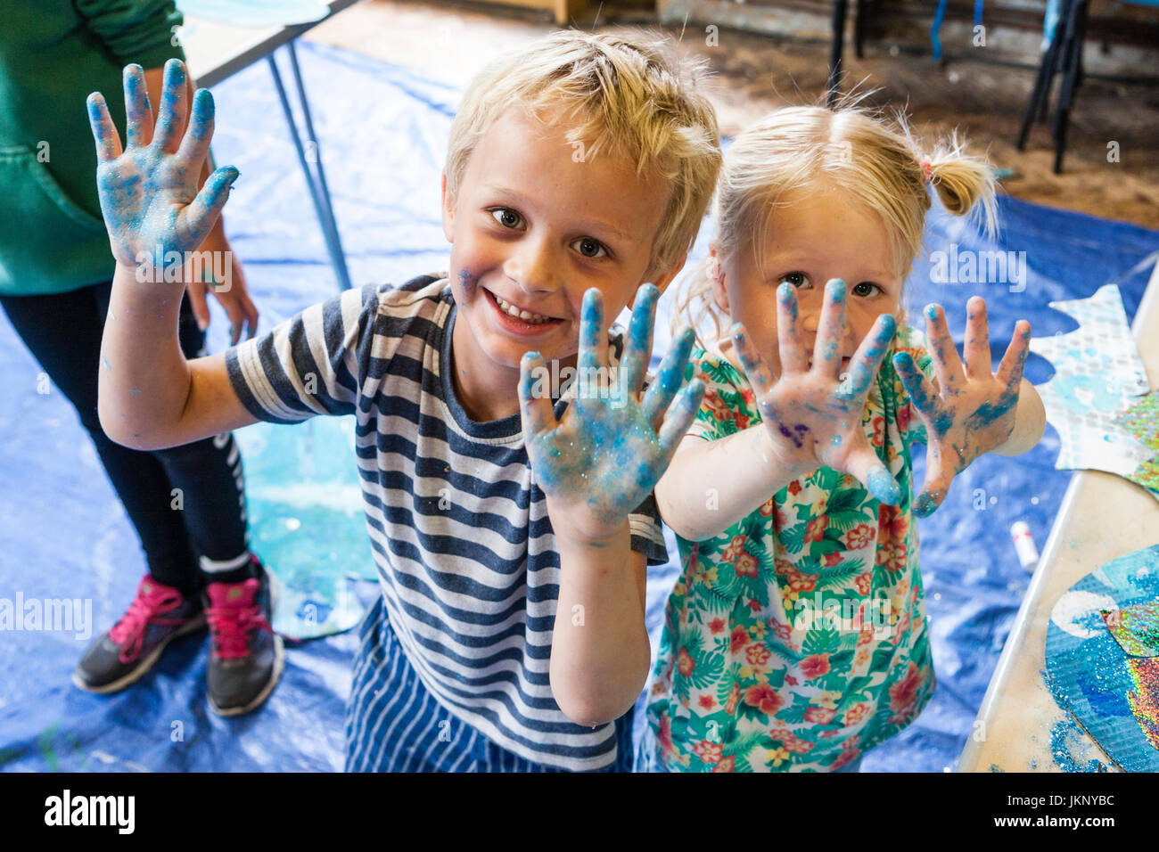 Smiling Caucasian blonde deux enfants, garçon, 5-6 ans, et une fille, 4-5 ans, frère et soeur, à la visualisation, au contact des yeux, montrant leurs mains peintes en désordre après un atelier d'art et d'artisanat. Banque D'Images