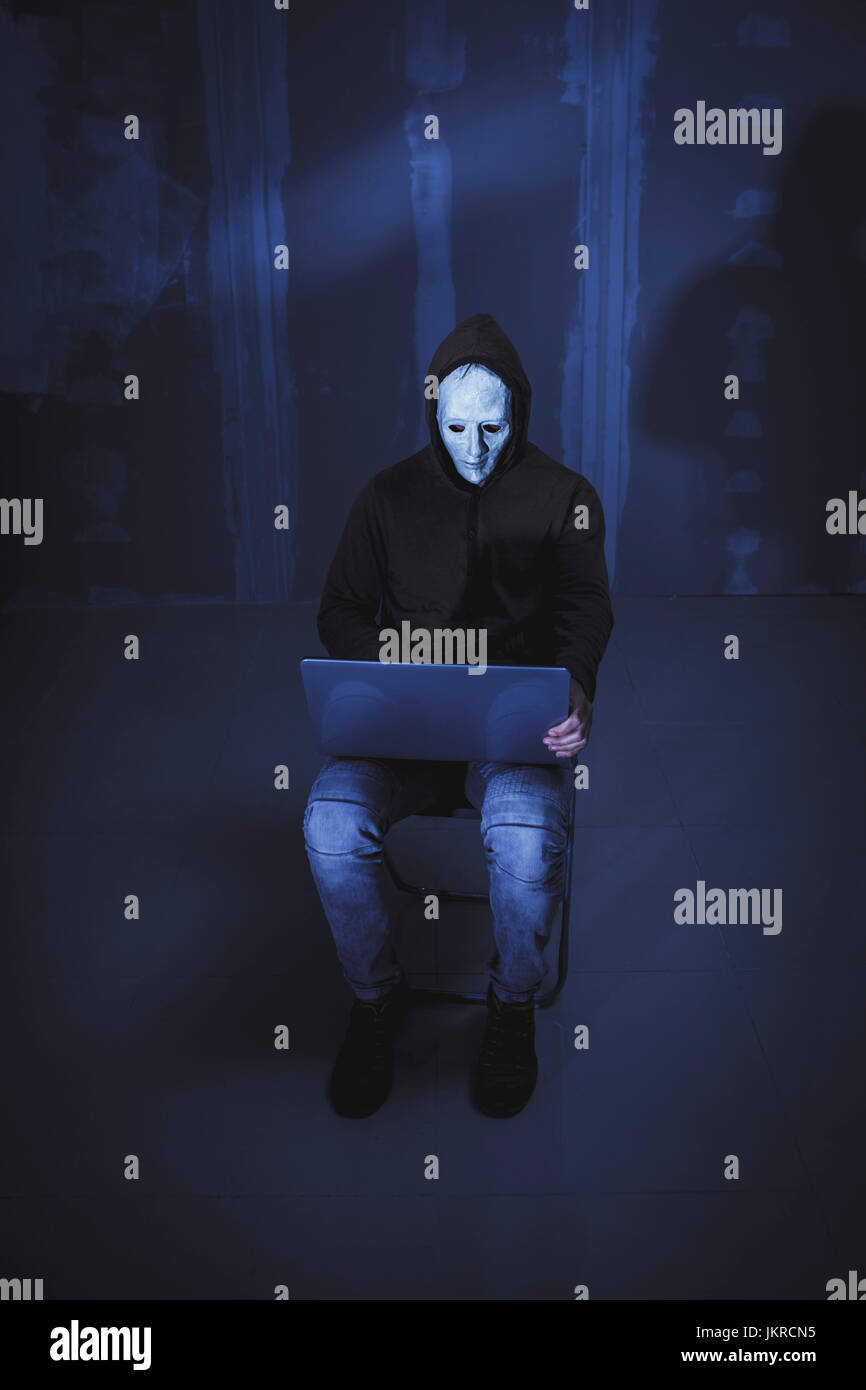 Toute la longueur du pirate informatique wearing mask et using laptop while sitting against wall Banque D'Images