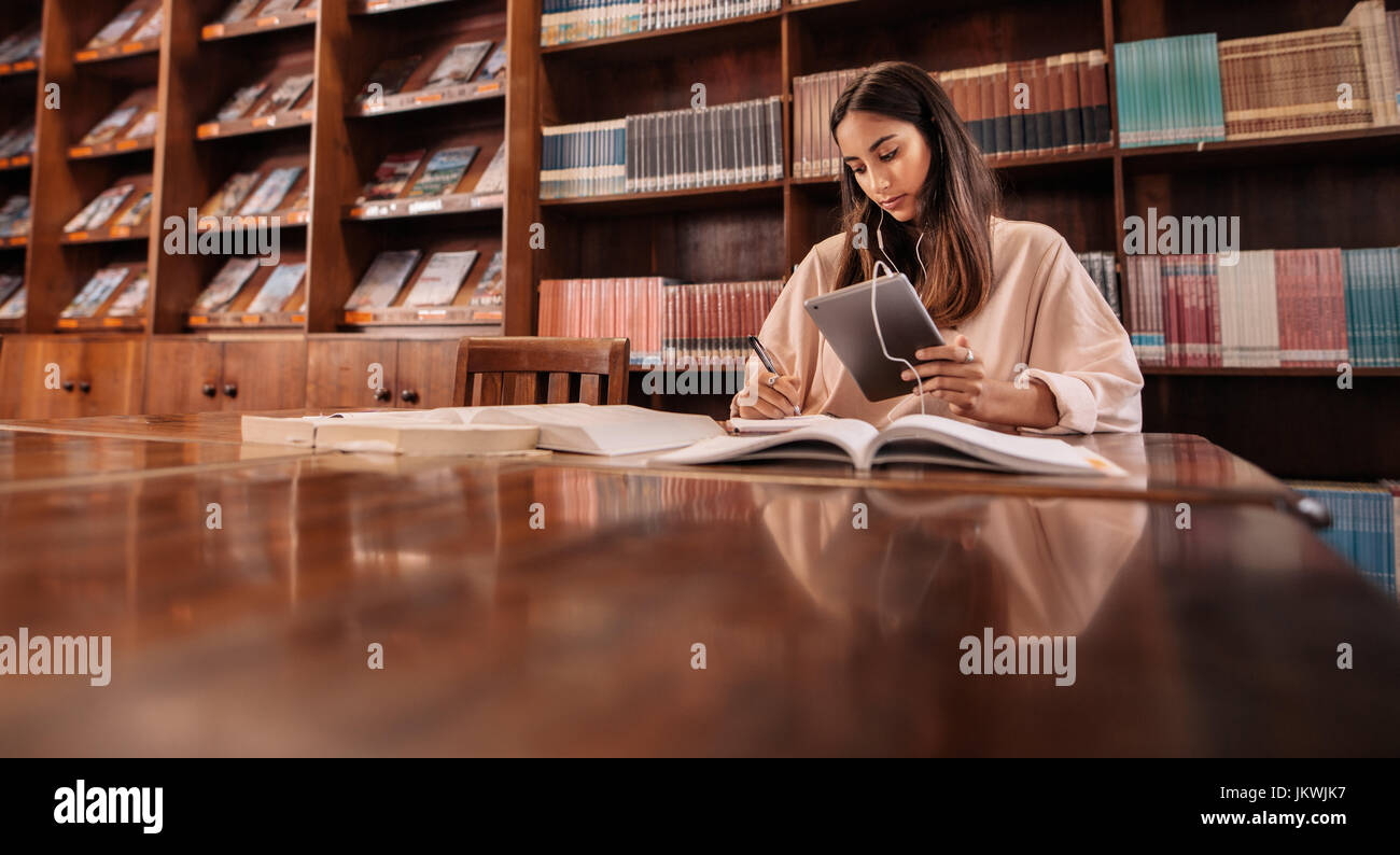 Beau jeune étudiant à l'aide de tablet pc et que la rédaction de notes dans la bibliothèque. College student taking notes de l'ouvrage de référence et d'écoute de musique Banque D'Images