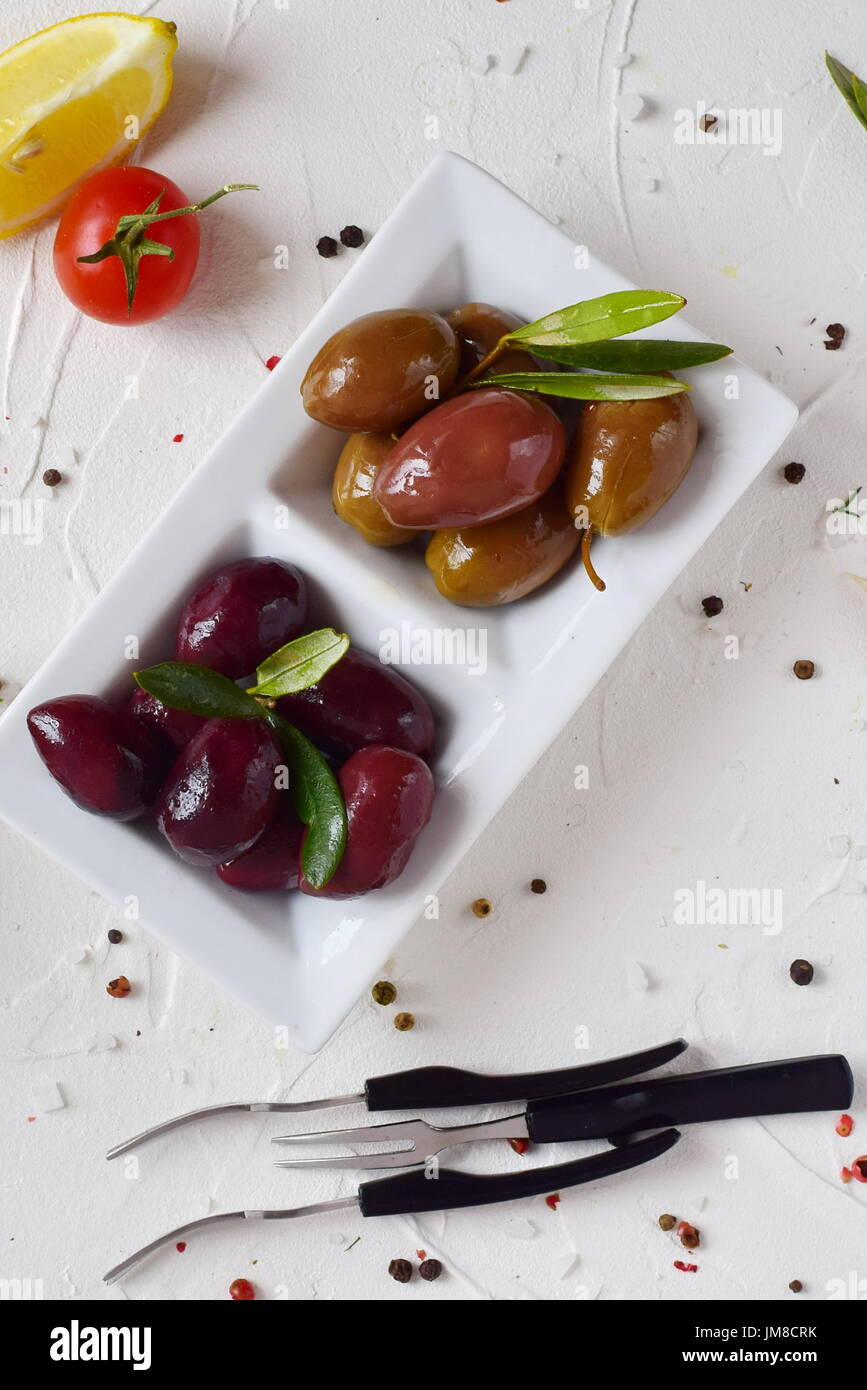 Plaque blanche avec olives vertes et noires avec feuilles d'olivier sur un fond abstrait blanc avec du citron, tomate cerise, d'épices. Alimentation saine. La nourriture grecque. La cuisine méditerranéenne. Banque D'Images