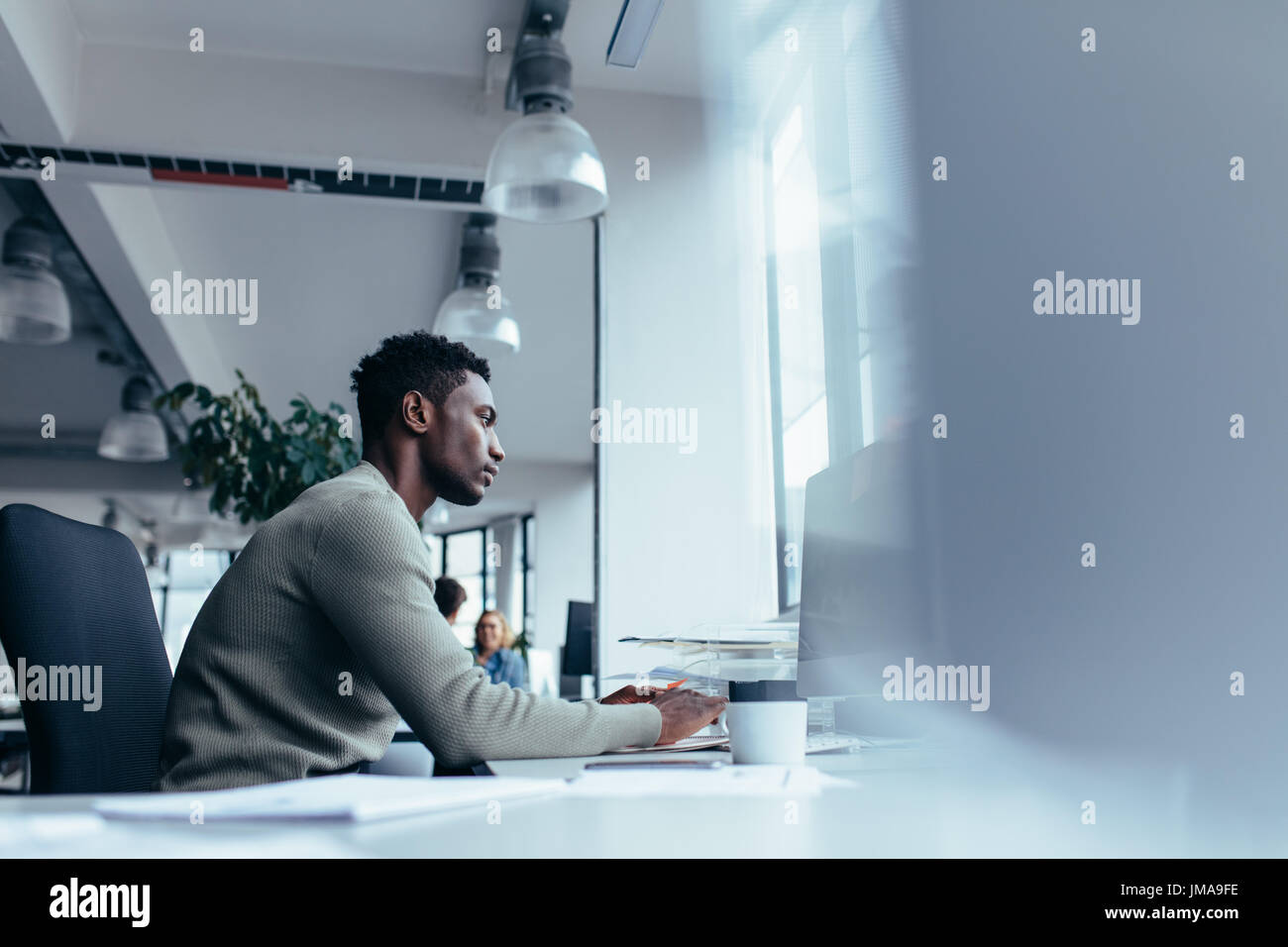 Side view of young African man working in office. Homme assis à son bureau exécutif et travaillant sur ordinateur de bureau. Banque D'Images