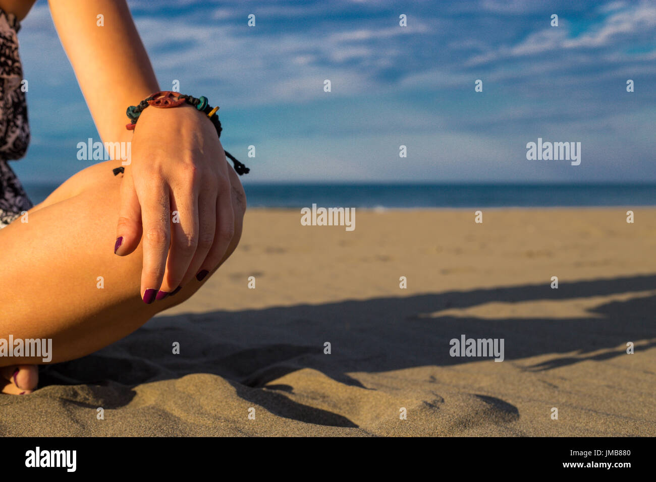 Femme assise sur la plage avec libre de tenir la main avec bracelet et de vernis à ongles sur le genou avec ombre, ciel bleu et la mer en arrière-plan summ Banque D'Images