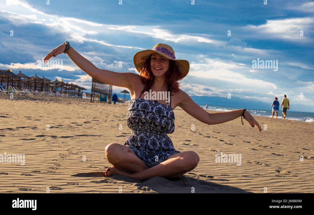 Happy happy summertime femme portant robe et chapeau assis sur la plage avec parasols et propagation d'armes et de ciel bleu et la mer en arrière-plan Banque D'Images