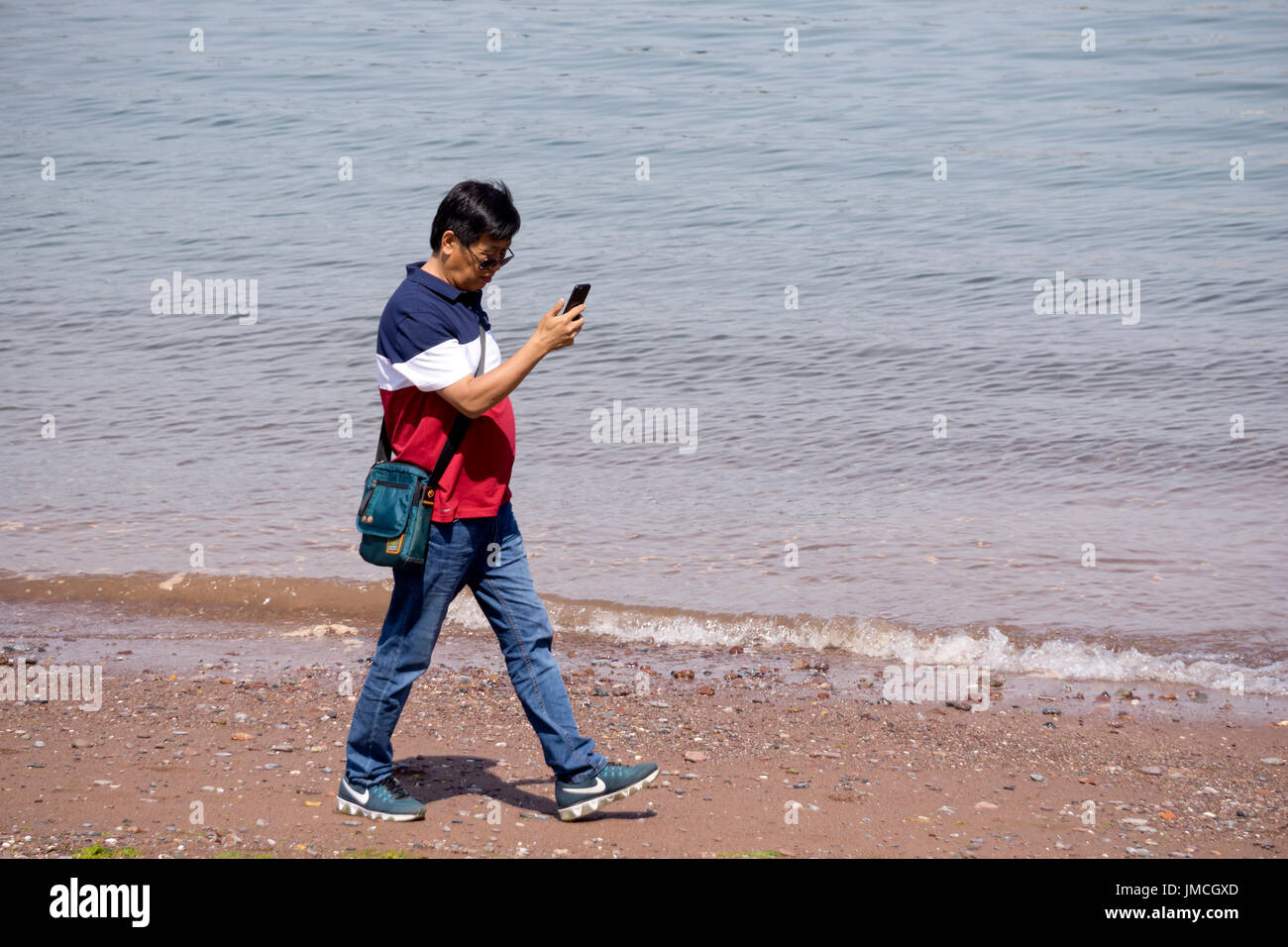 Homme marchant le long de la plage en regardant son téléphone Banque D'Images