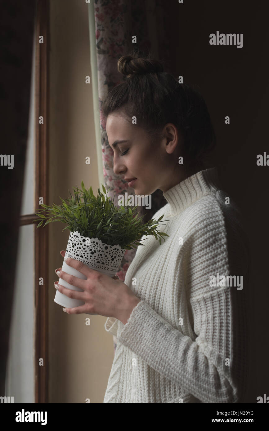 Vue latérale du smiling woman smelling potted plant en position debout par fenêtre à la maison Banque D'Images Vue latérale du smiling woman smelling potted plant en position debout par fenêtre à la maison Banque D'Images