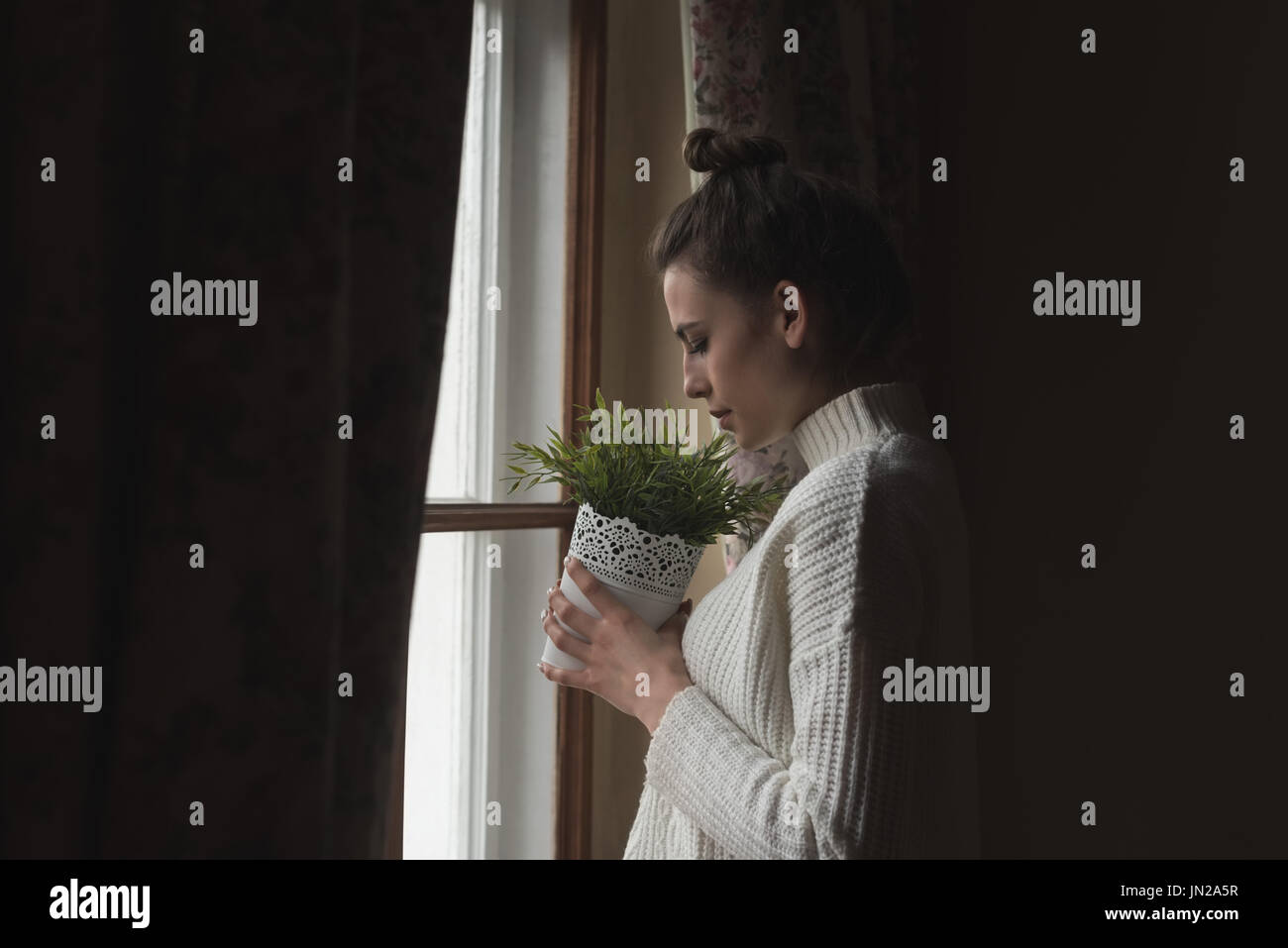 Vue latérale du woman holding potted plant en position debout par fenêtre à la maison Banque D'Images Vue latérale du woman holding potted plant en position debout par fenêtre à la maison Banque D'Images