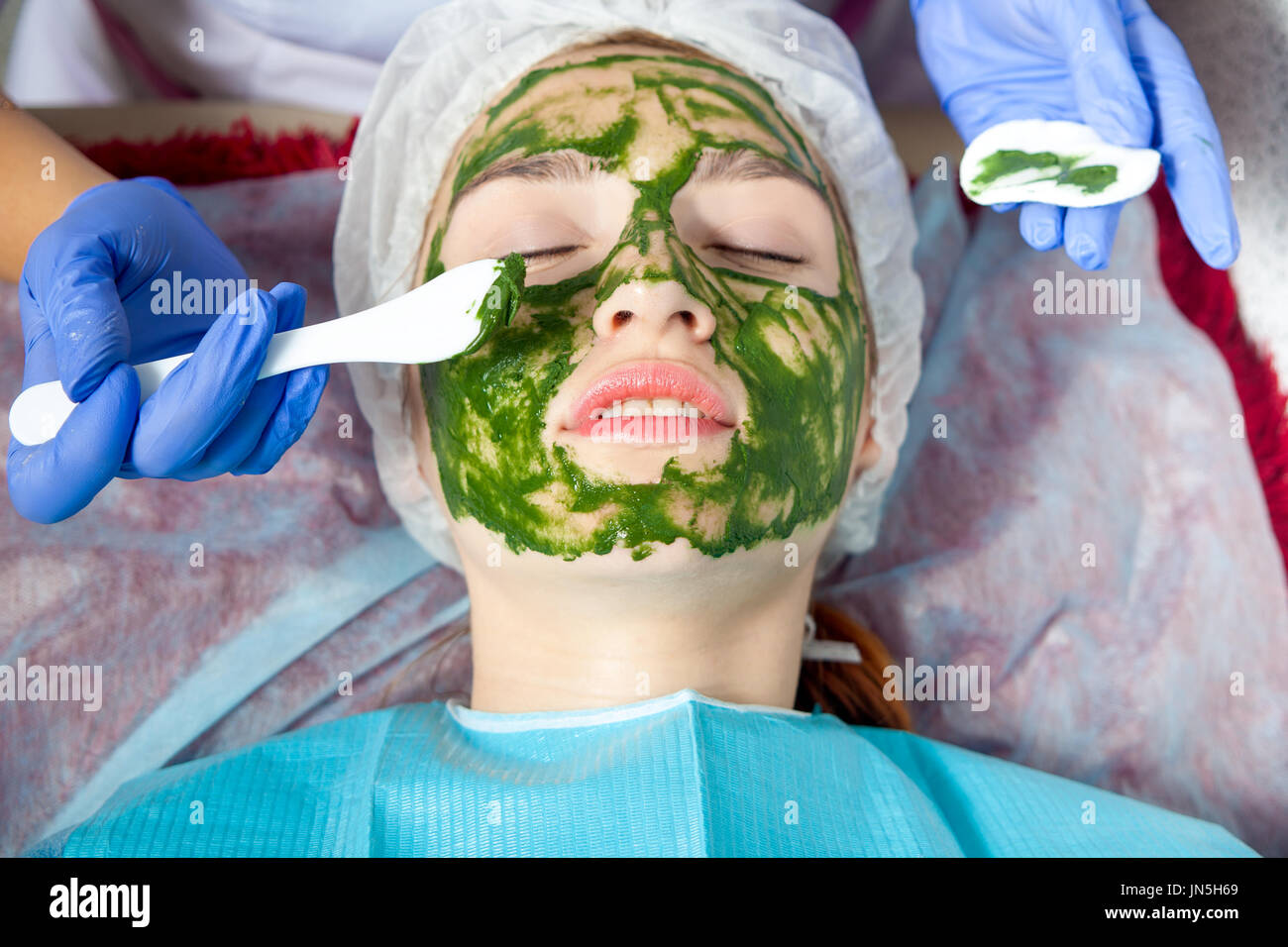 Un gros plan de l'esthéticienne en bleu à l'aide de gants spéciaux un pinceau de maquillage fait un masque de spa thérapeutiques d'algues bleues contre le vieillissement d'un jeune h-rouge Banque D'Images