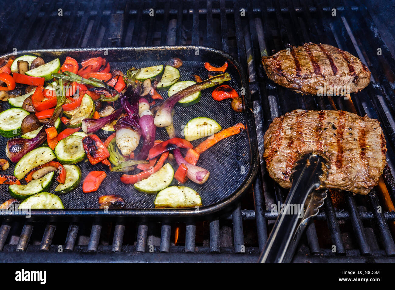 Deux steaks étant grillées sur le barbecue avec des légumes grillés. Les légumes : courgettes, poivron rouge, oignon vert, champignons et Banque D'Images