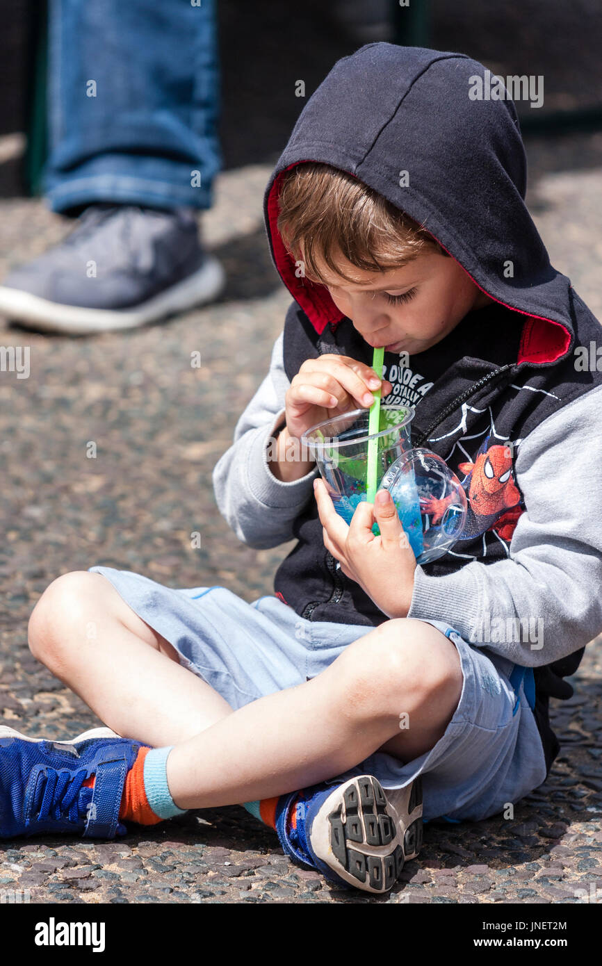 Caucasien enfant, garçon, 5-6 ans, assis en tailleur dans la rue en plein soleil de boire avec une paille de plastique. Le port de shorts et de la hotte. Banque D'Images