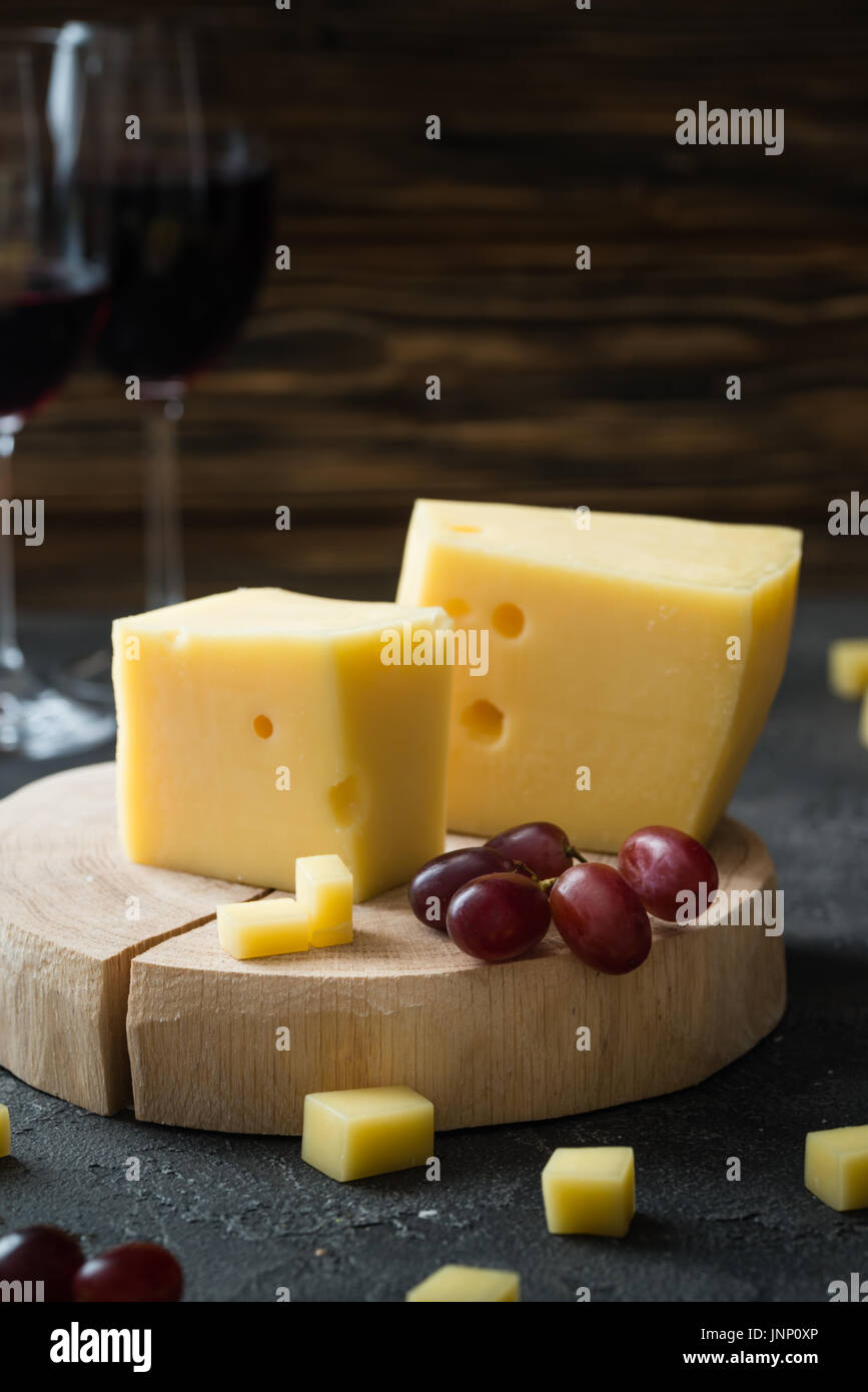 Disque suédois fromage jaune avec des trous coupé avec raisins rouges sur tranches de bois et de verres de vin rouge foncé sur fond rustique Banque D'Images