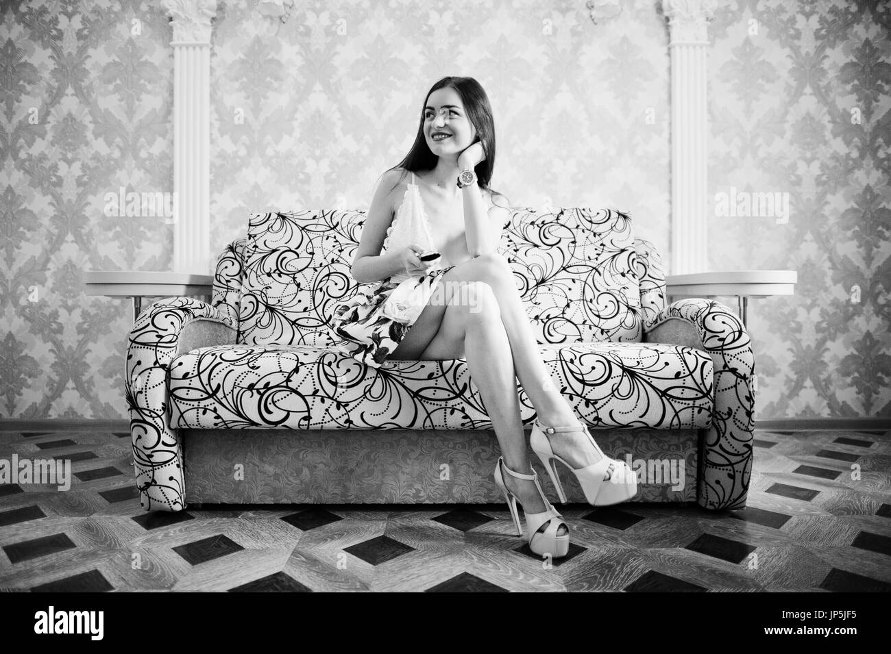 Portrait d'une belle jeune fille en robe à fleurs assis sur le canapé avec une télécommande dans les mains. Photo en noir et blanc. Banque D'Images