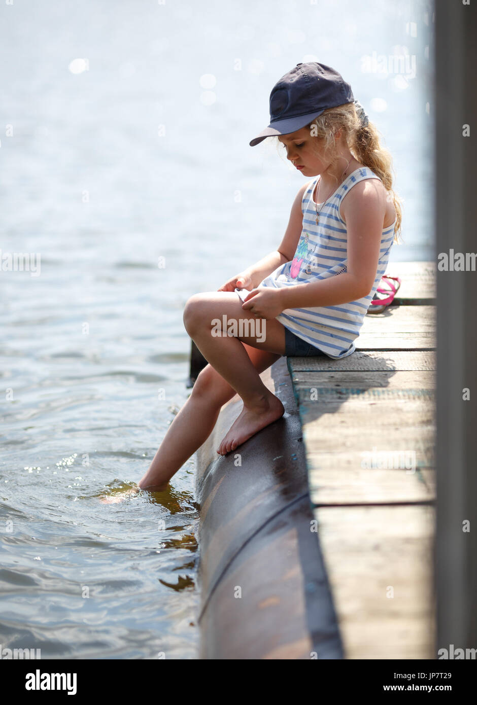 Petite fille assise sur une plate-forme de bois par la rivière ou du lac tandis que les projections d'eau par les pieds. Banque D'Images