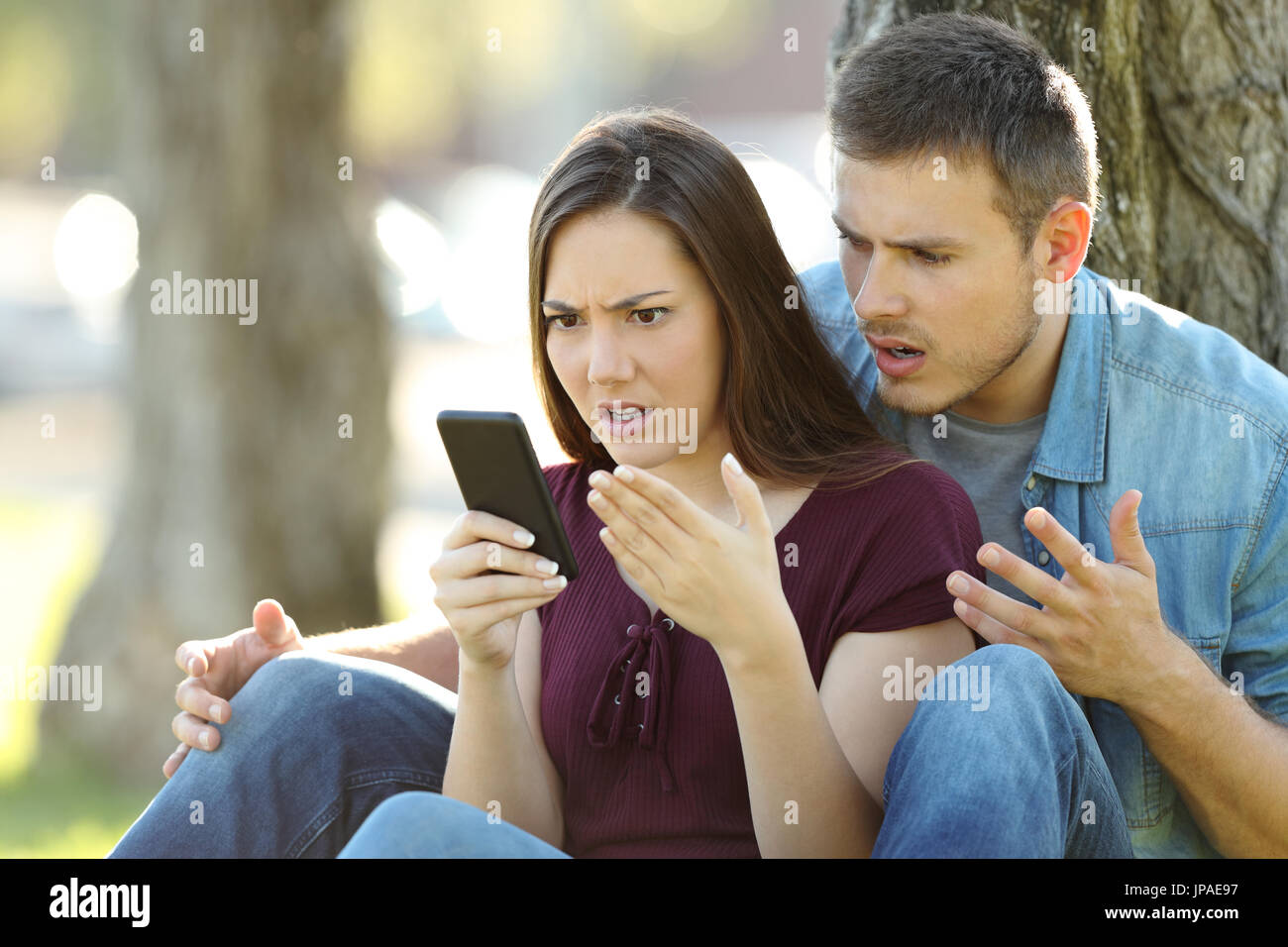 Angry couple à l'aide d'un smart phone outdoors sitting in a park Banque D'Images