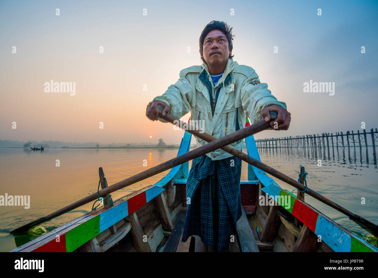 Amarapura, région de Mandalay, Myanmar. L'homme de l'aviron sur son bateau coloré sur le lac Taungthaman au lever du soleil, avec l'U Bein bridge en arrière-plan. Banque D'Images