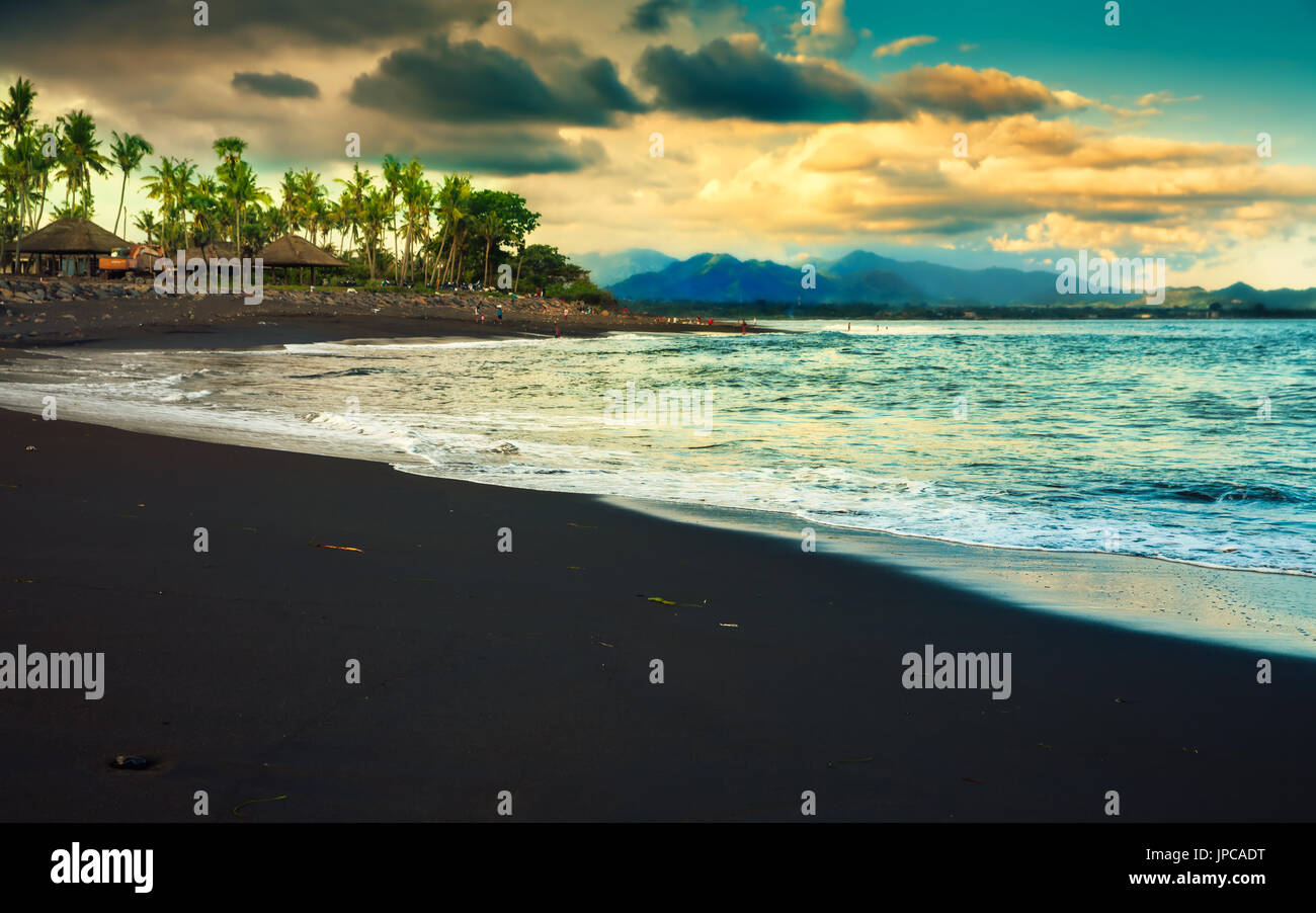 Plage volcanique de sable noir sur fond de nature tropicale étonnante avec green palm tree et les paysages de montagne à l'horizon au coucher du soleil. L'Indonésie Banque D'Images