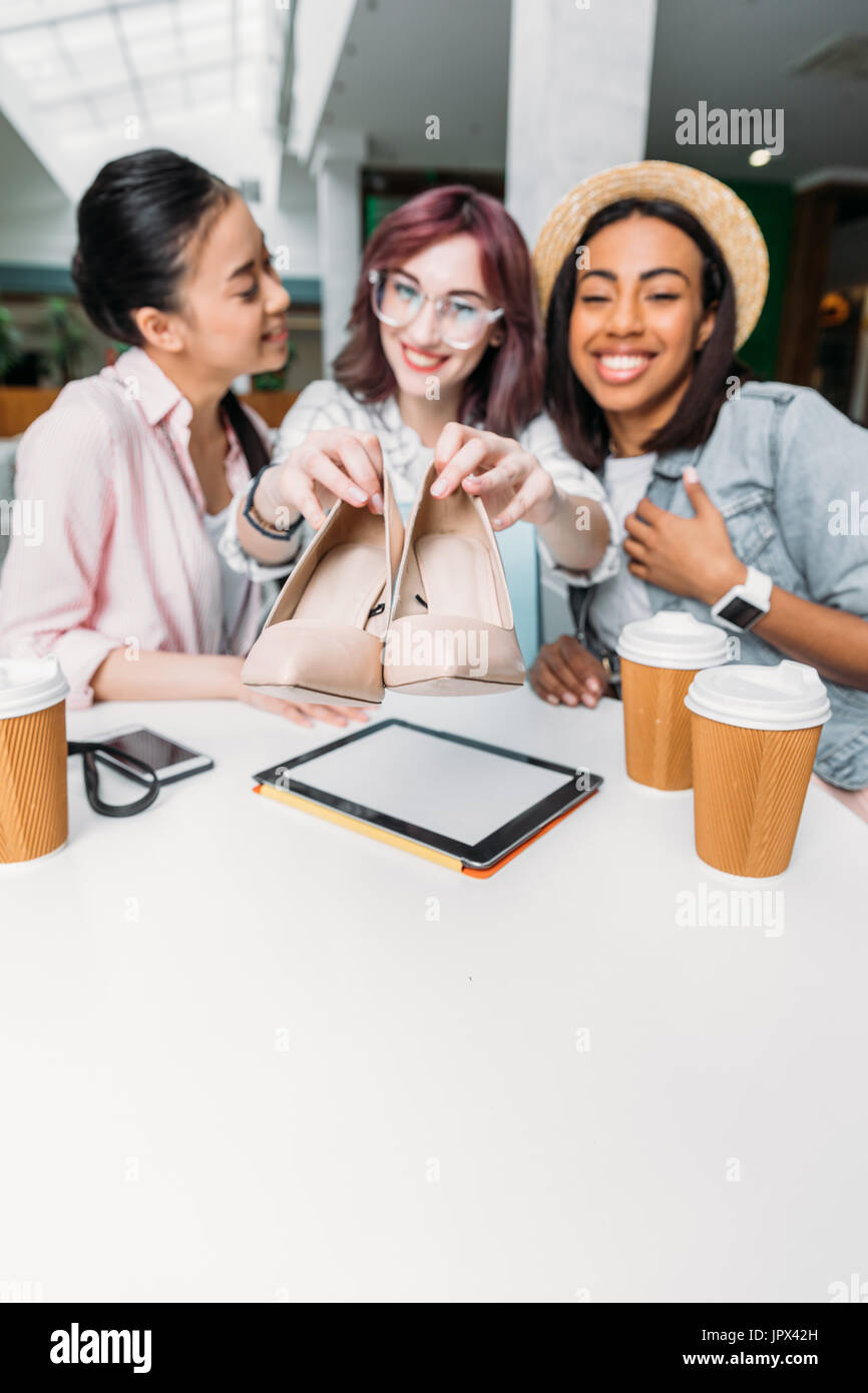 Souriante jeune femme assis à table avec les tasses de papier et montrant les nouvelles chaussures élégantes, les jeunes filles shopping concept Banque D'Images