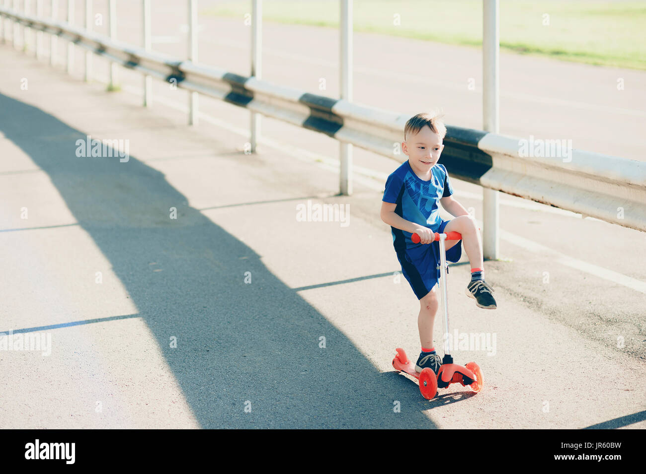 Petit garçon sur un scooter dans un parc de la ville, à l'extérieur. Dans un costume d'été bleu repéré, sur un scooter orange, il roule une jambe sur le volant Banque D'Images