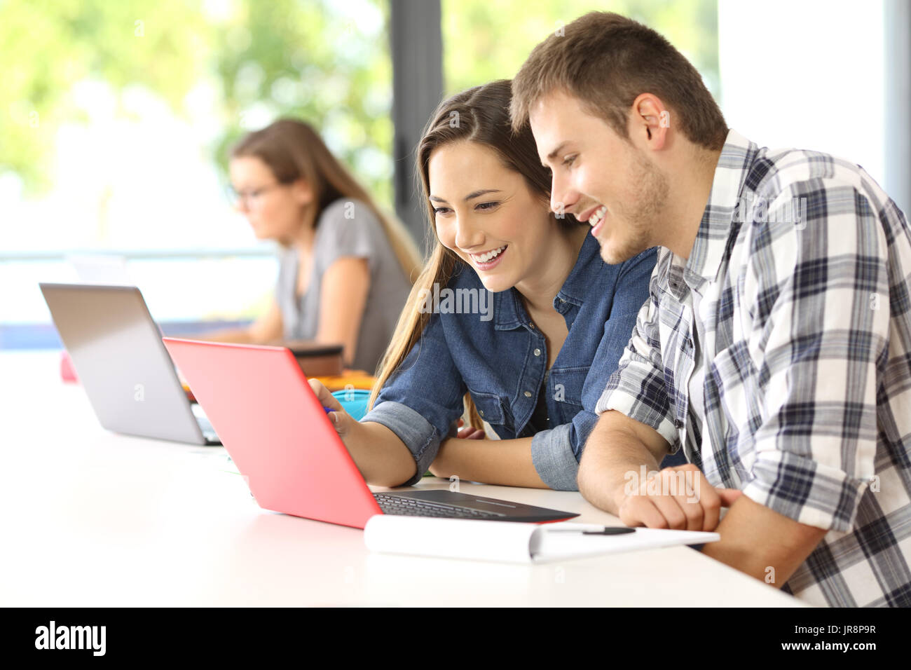 Deux étudiants heureux sur la ligne d'apprentissage avec un ordinateur dans une classe avec d'autres camarades dans l'arrière-plan Banque D'Images