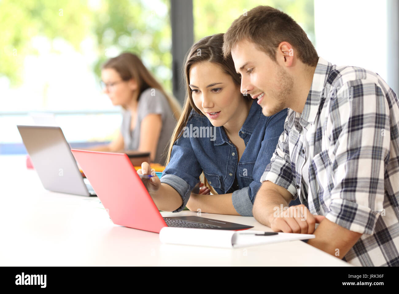 Deux étudiants concentrés sur ligne, assis dans un bureau dans une salle de classe avec un camarade dans l'arrière-plan Banque D'Images